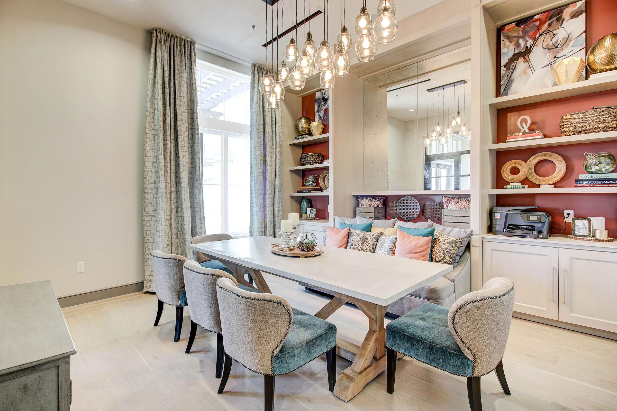 A modern dining area featuring a light wood table surrounded by six upholstered chairs. In the background, there are built-in shelves decorated with various ornaments, cushions, and a printer. Large windows allow natural light to fill the space, complemented by stylish pendant lighting overhead.