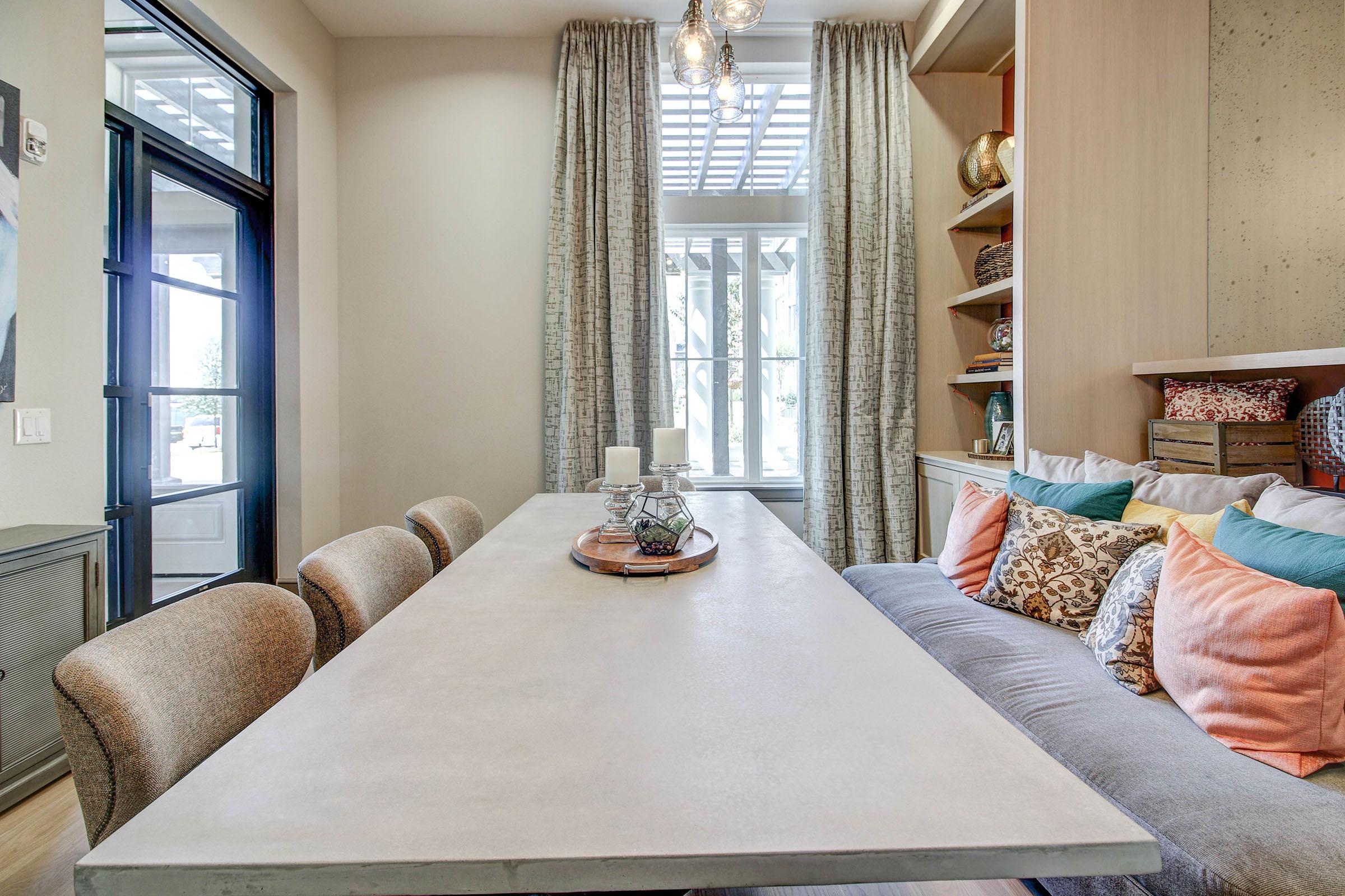 A modern dining area featuring a large concrete table surrounded by upholstered chairs. A cozy sofa with colorful pillows is positioned against the wall, with shelves showing decorative items. Natural light flows in through large windows, complemented by elegant curtains and stylish light fixtures overhead.