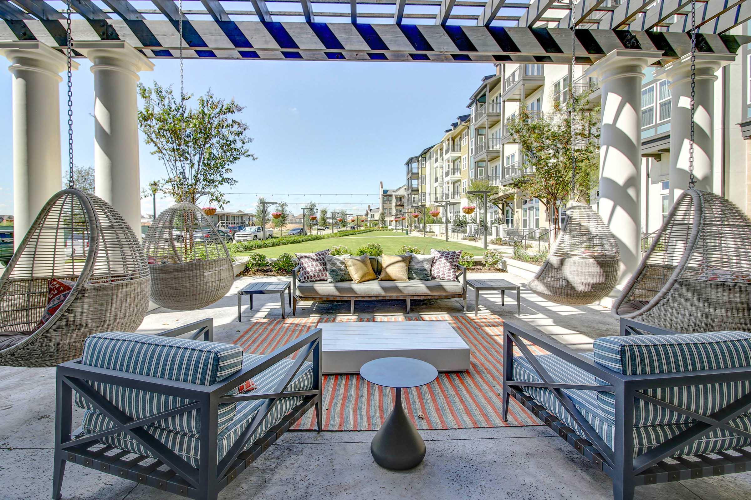 A stylish outdoor lounge area featuring hanging wicker chairs, a cozy sofa with colorful cushions, and modern furniture. The space is surrounded by green landscaping, with a view of nearby buildings and a clear blue sky, creating a vibrant and inviting atmosphere.