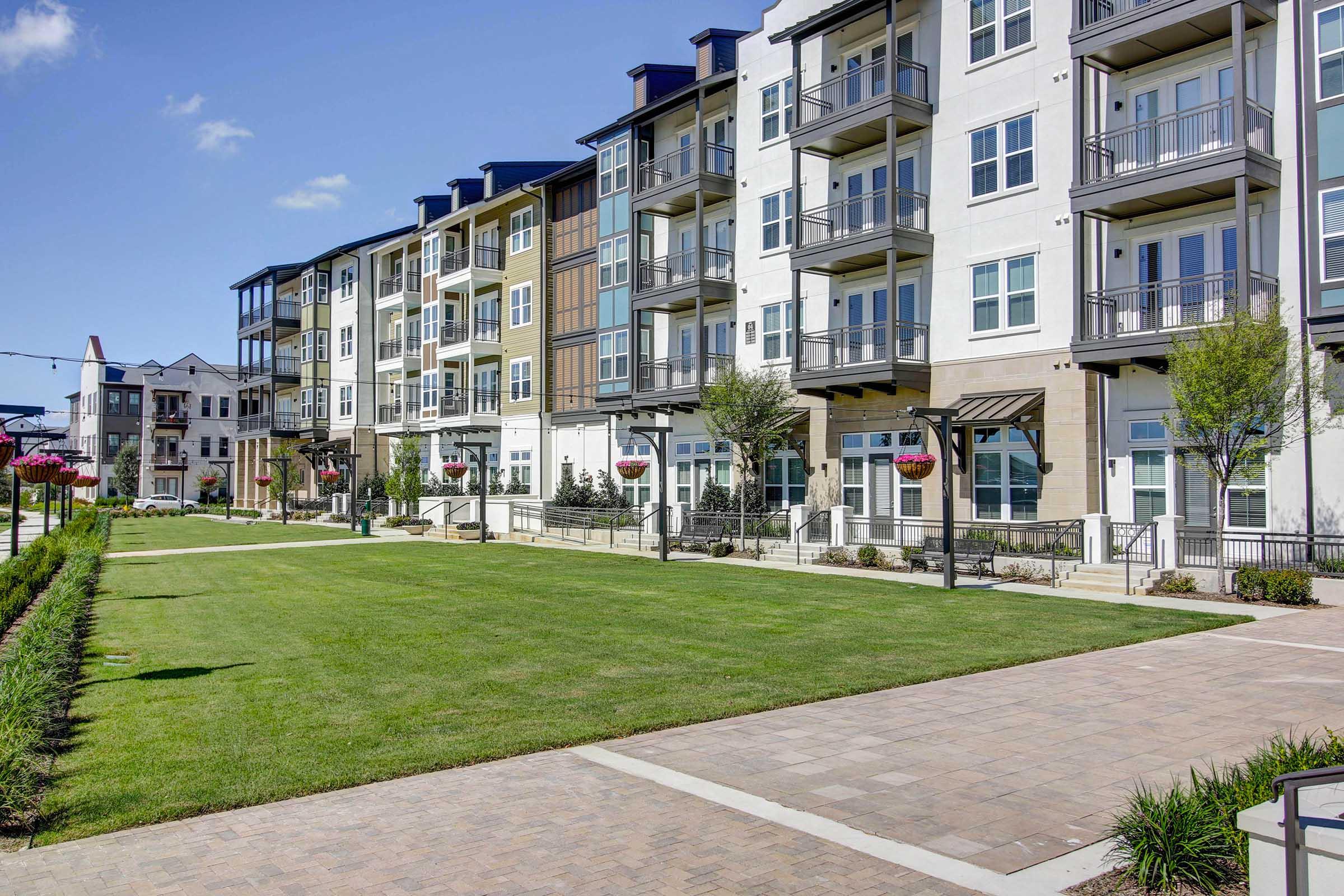 A modern apartment complex featuring three-story buildings with balconies, large windows, and a well-maintained green lawn. There are flower planters along a paved walkway, adding a touch of color, with clear blue skies above. The area appears inviting and well-designed for residential living.