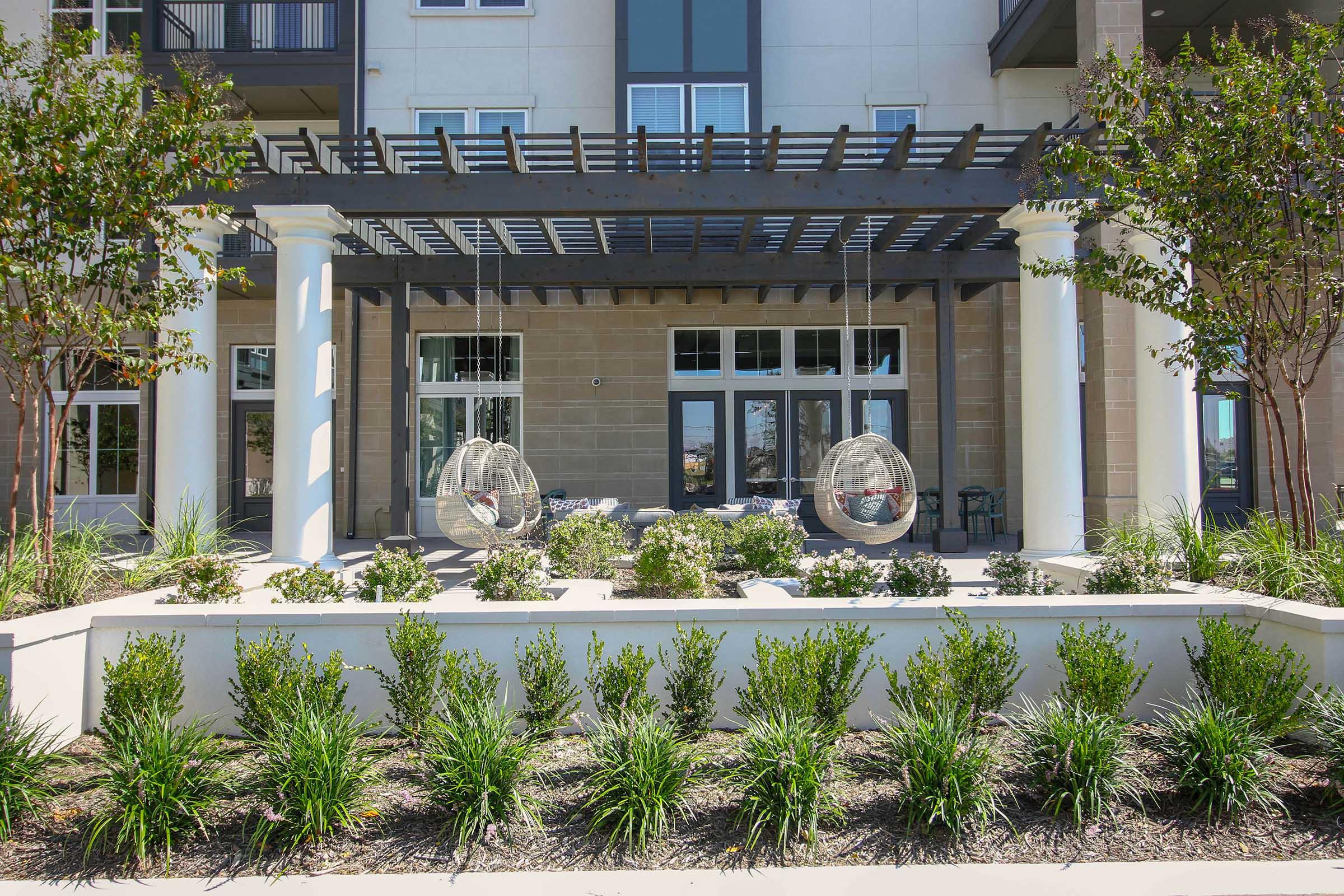 A modern outdoor patio area featuring two hanging chairs suspended from a pergola. In the foreground, neatly arranged greenery and low shrubs enhance the landscaping. The backdrop includes a building facade with large windows and doors, creating an inviting atmosphere.