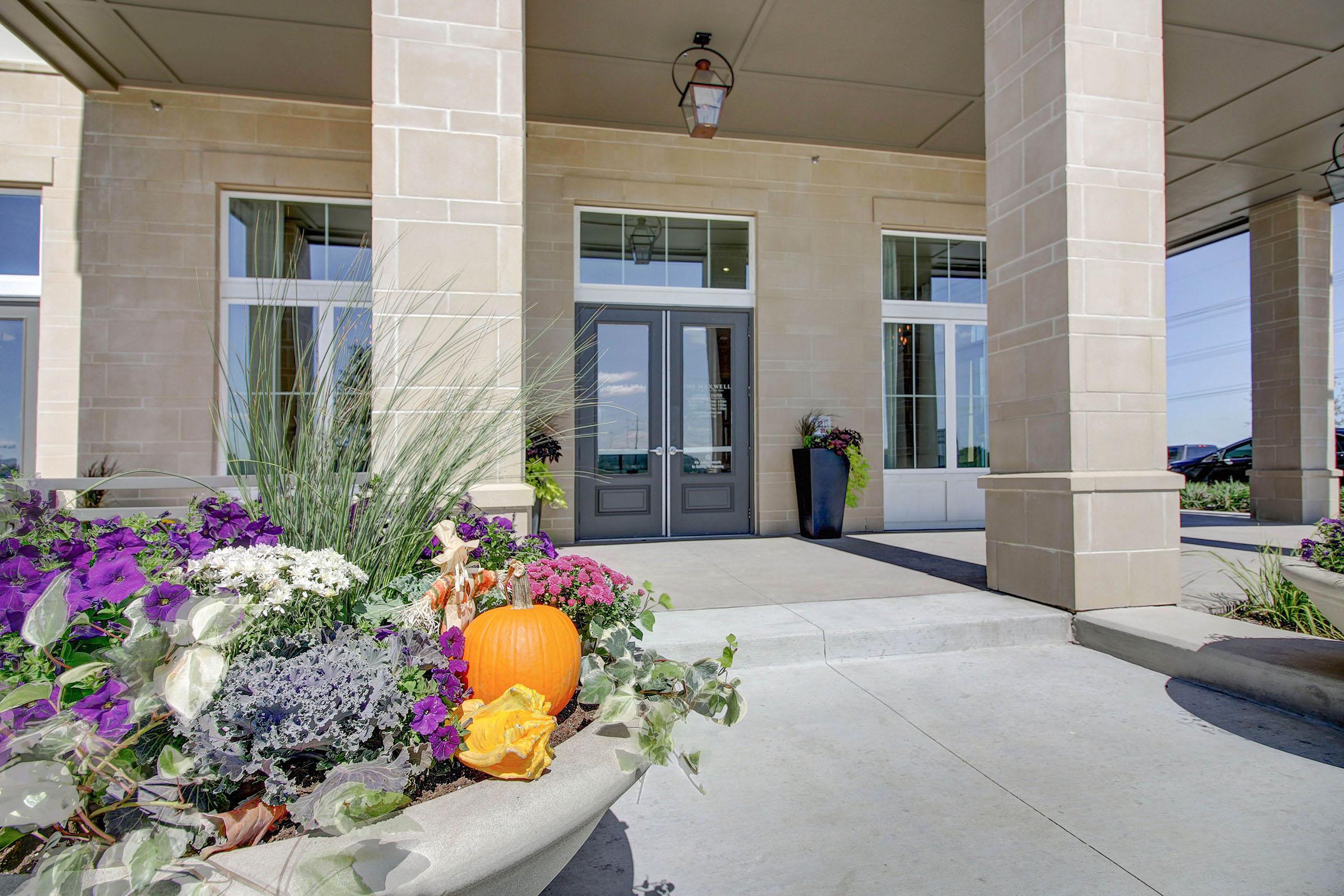 A welcoming entrance featuring large stone pillars, double doors in a muted gray color, and a vibrant flower bed with purple and white blooms. A small orange pumpkin adds a seasonal touch, enhancing the inviting atmosphere of the building's facade.