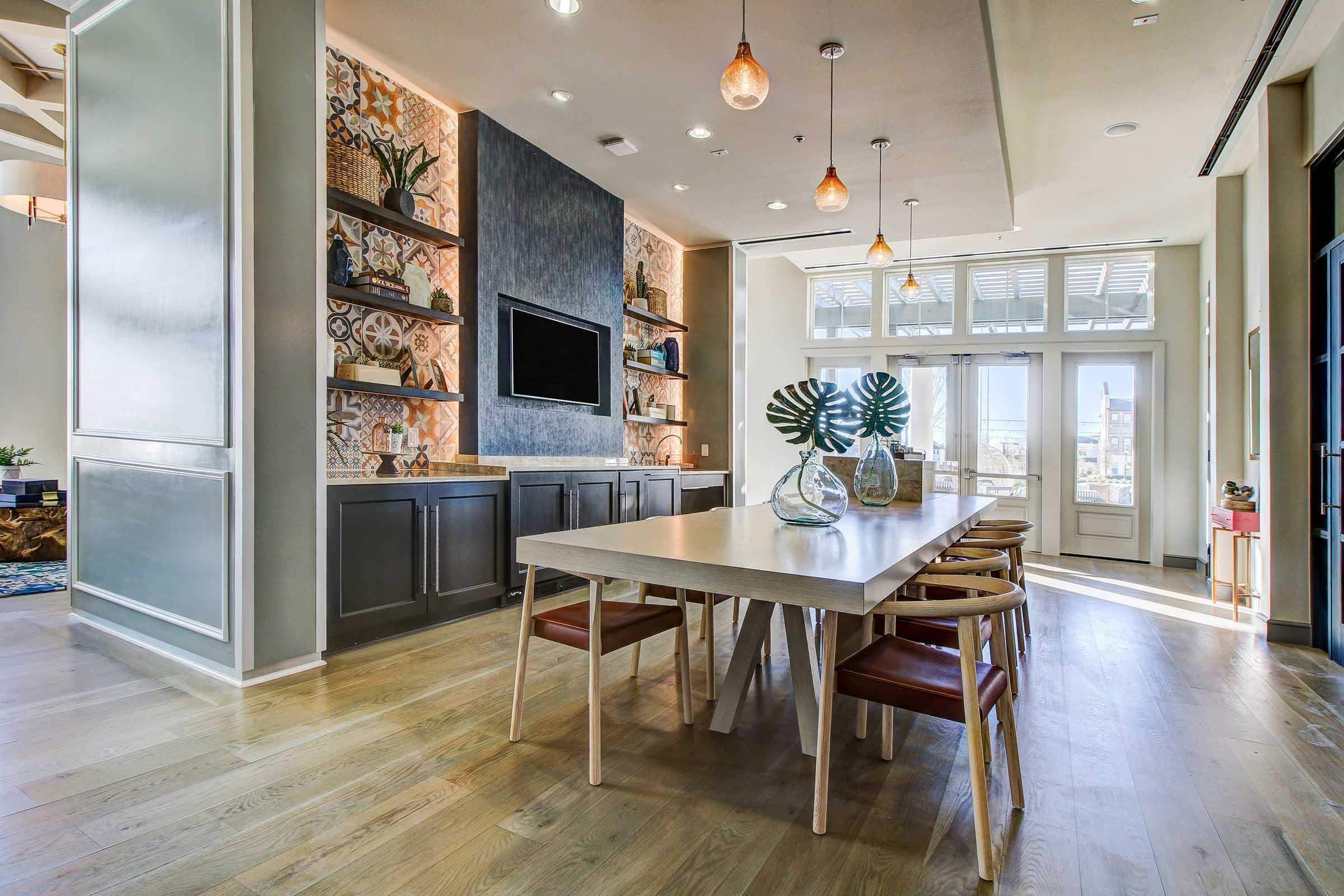 A modern dining area featuring a large rectangular table surrounded by wooden chairs. The walls are adorned with decorative shelves and an accent wall with a textured design. Natural light floods in through large windows, complementing the minimalist decor and warm wood flooring.