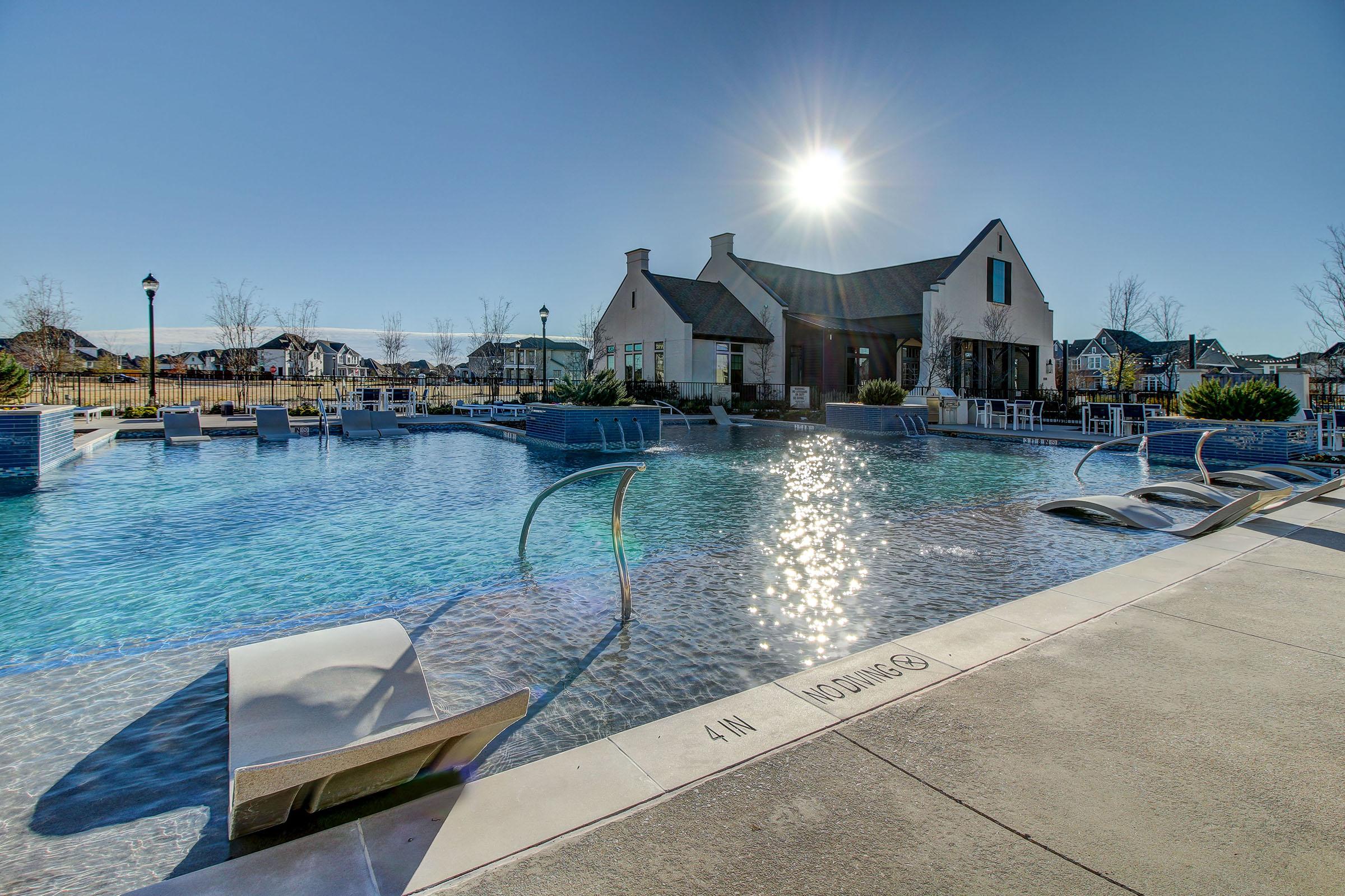 A tranquil outdoor swimming pool surrounded by modern lounge chairs and a clubhouse. The sun shines brightly in a clear blue sky, casting reflections on the water's surface. The setting is peaceful, with landscaped greenery in the background, and a clear indication of water depth marked on the poolside.