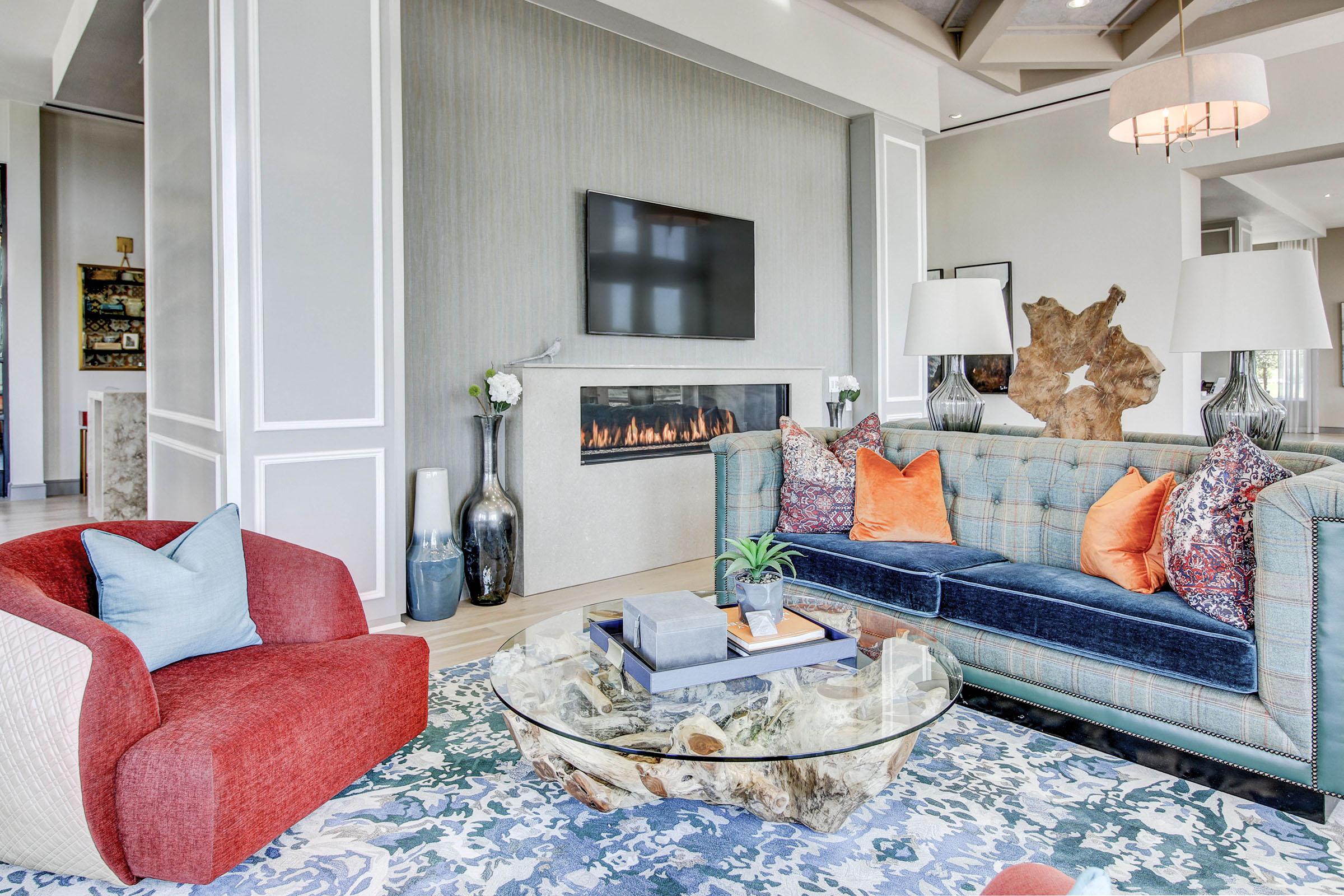 A modern living room featuring a blue and red sofa with colorful throw pillows, a round glass coffee table on a patterned rug, elegant lamps, and a contemporary fireplace under a wall-mounted television. Bright, light-filled space with decorative plants and artwork in the background.