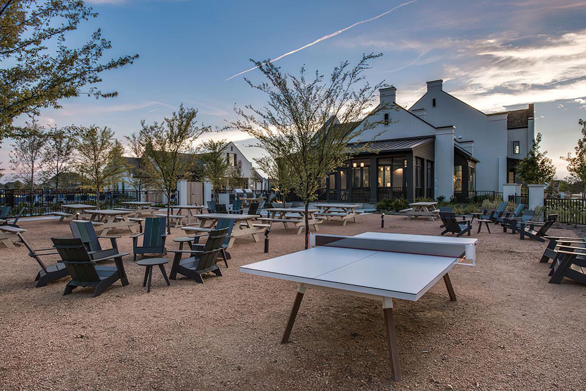 A landscaped outdoor area featuring several chairs and tables, with a ping pong table prominently in the foreground. Trees are scattered throughout the space, and a modern building can be seen in the background under a cloudy sky at dusk.