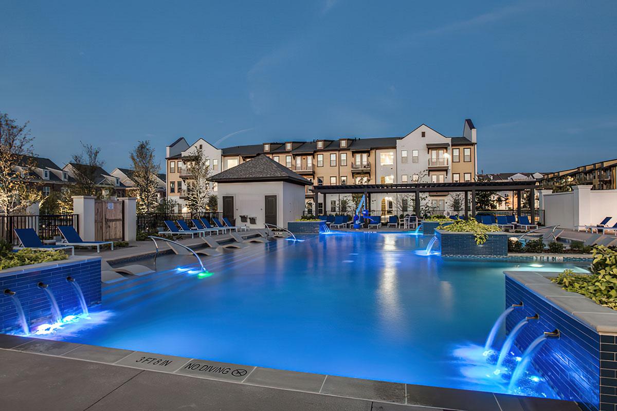 A tranquil outdoor swimming pool at dusk, featuring illuminated water features and lounge chairs. In the background, modern apartment buildings are visible along with greenery and paved areas, creating a relaxing atmosphere. The scene is enhanced by soft lighting and a clear sky.
