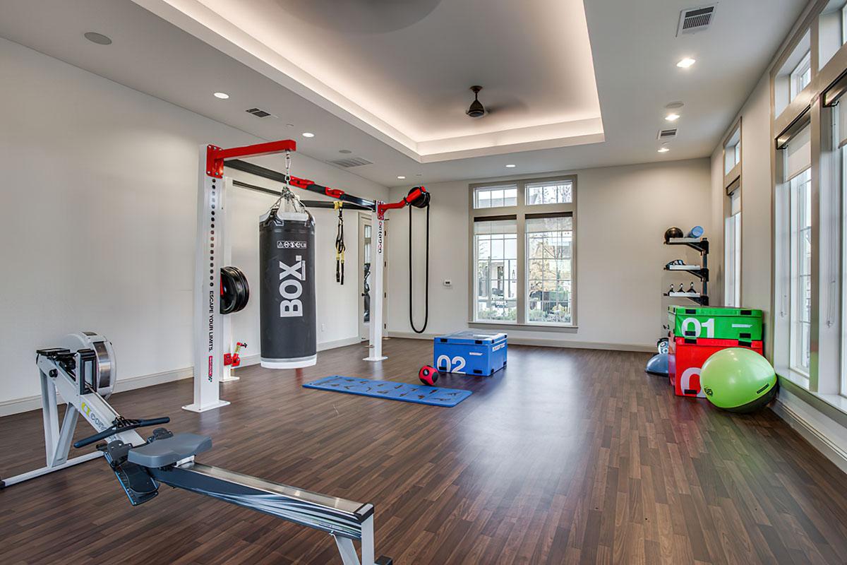 A modern home gym featuring a boxing bag, rowing machine, exercise mats, and various fitness equipment. Large windows allow natural light, and the wooden floor provides a warm atmosphere. Colorful storage boxes and exercise balls are arranged for easy access.