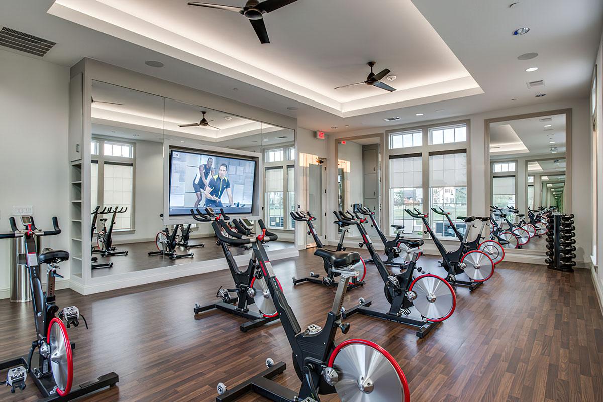 A modern indoor cycling studio featuring several stationary bikes arranged in rows, large mirrors on the walls, and a presentation screen displaying a fitness class. The room is brightly lit with wooden flooring and large windows offering natural light.