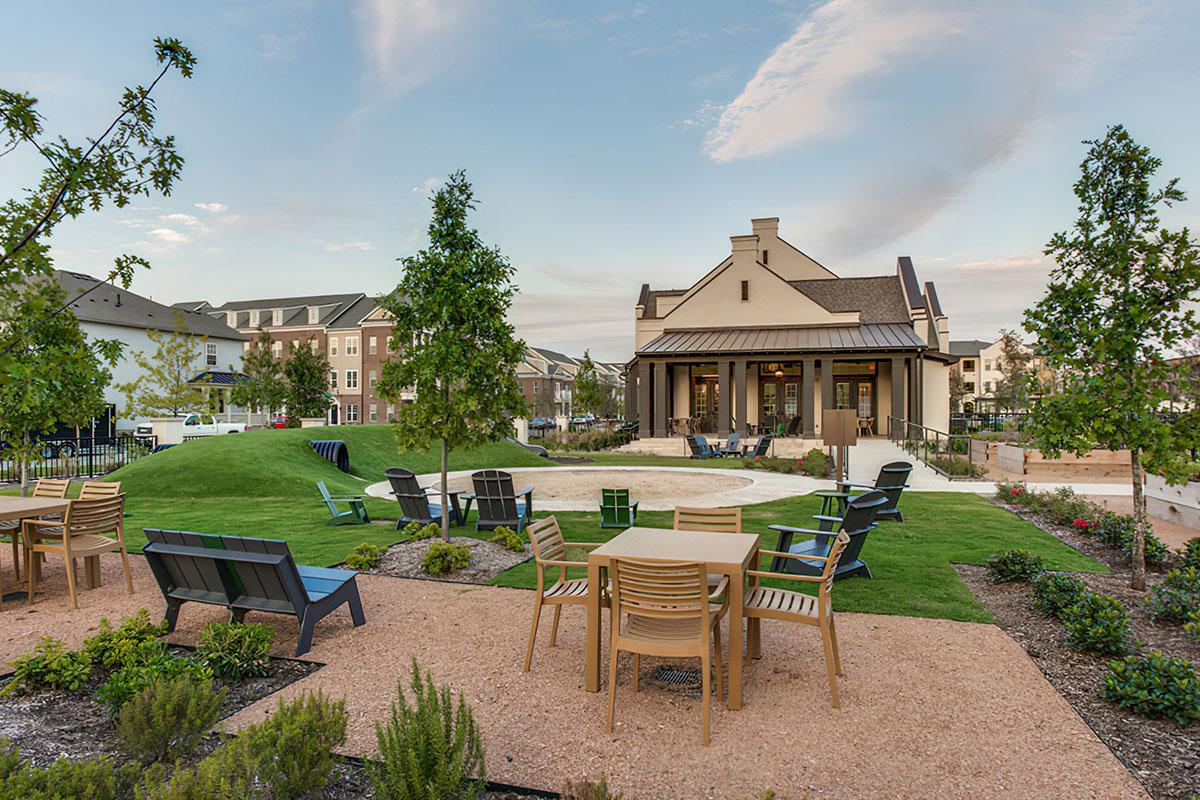 A landscaped outdoor area featuring several tables and chairs, surrounded by greenery and trees. In the background, a modern building serves as a community space, with benches scattered around the grass. The sky is clear with soft clouds, creating a serene and inviting atmosphere.
