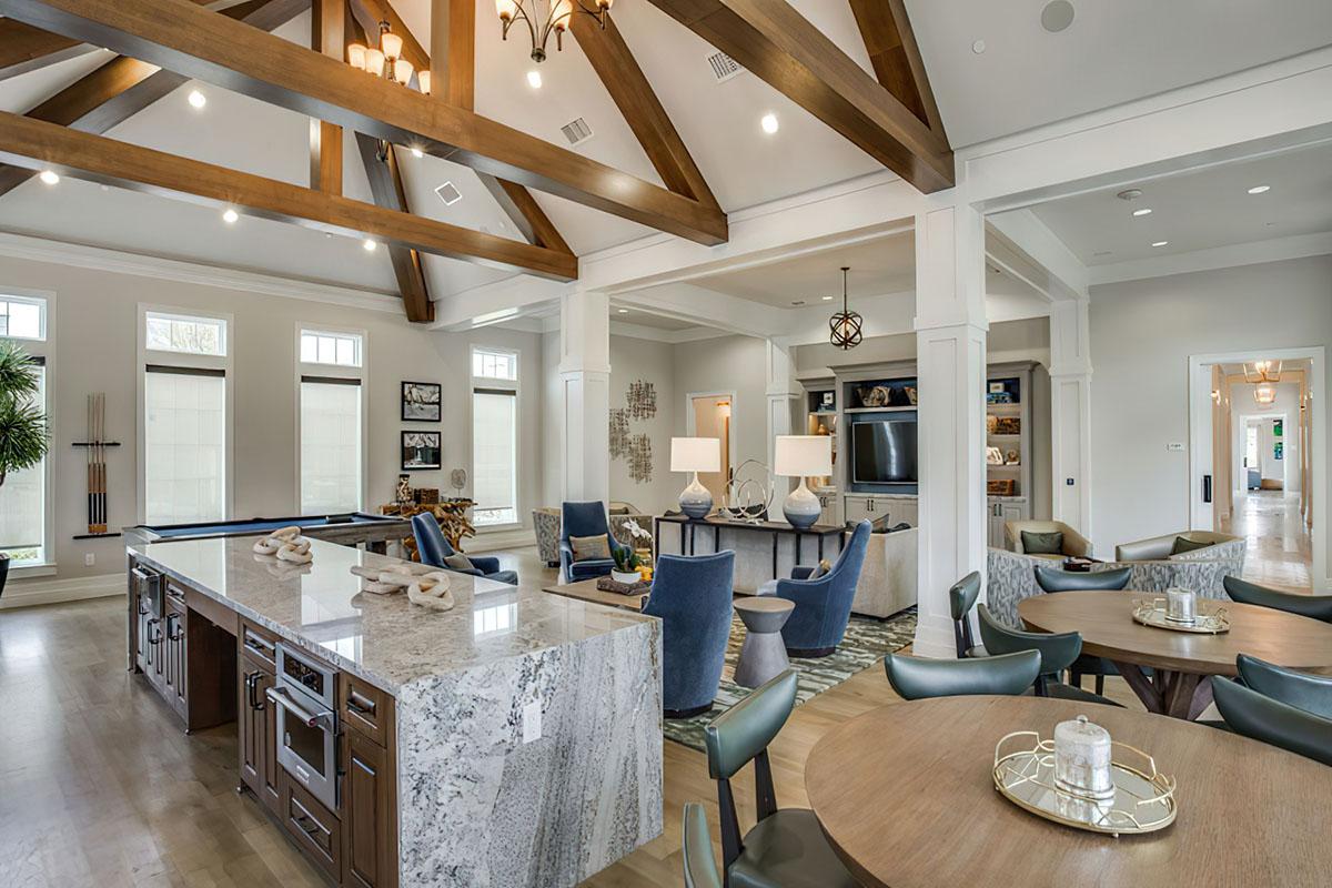 A spacious, modern living area featuring a large kitchen island with a marble countertop, surrounded by dark wood cabinets. The room is illuminated by stylish lighting and boasts high wooden beams on the ceiling. Comfortable seating areas include blue armchairs and round dining tables, with large windows allowing natural light.