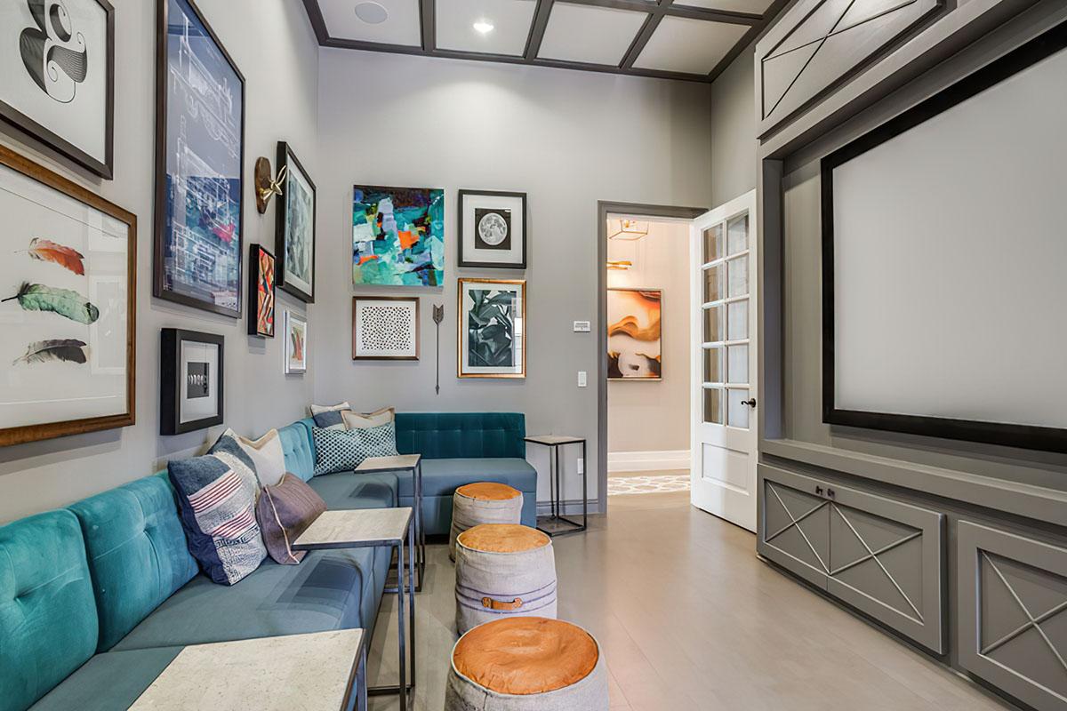 A modern living room featuring a teal bench with decorative cushions, round stools, and an accent wall adorned with various framed artworks. The room has a neutral color palette, elegant trim, and a large wall-mounted screen, with a doorway leading to another space.