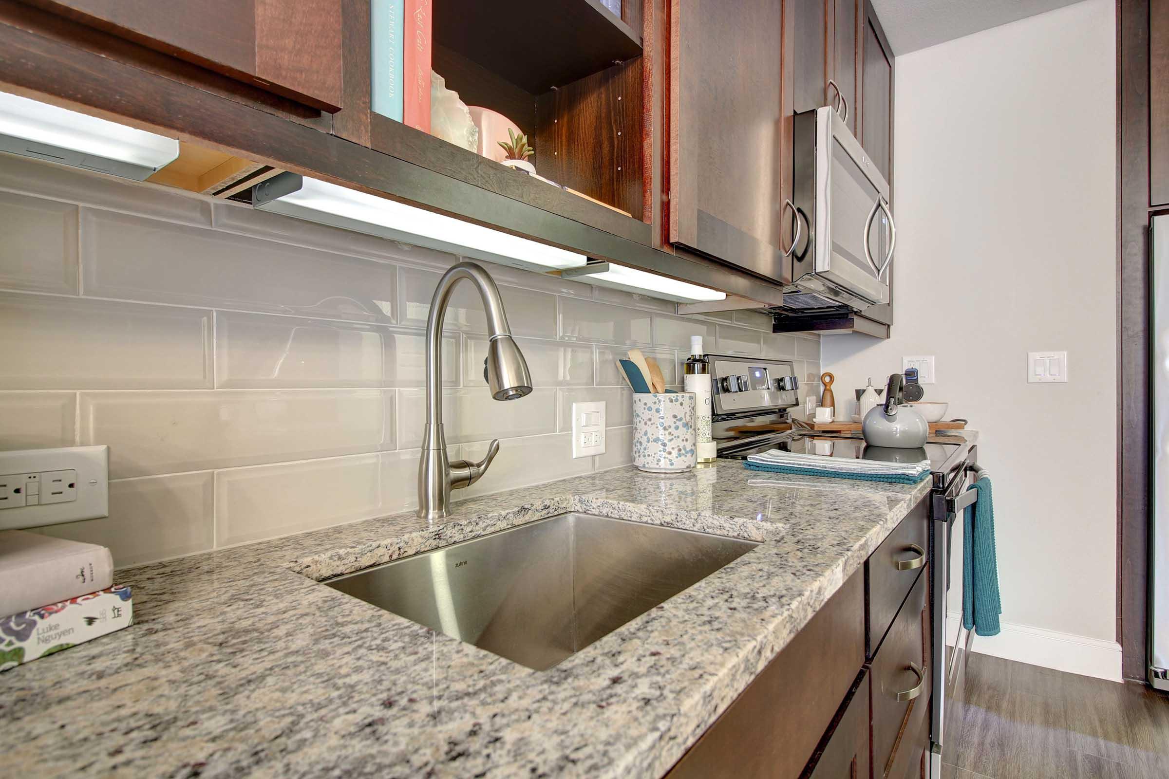 A modern kitchen featuring a stainless steel sink with a sleek faucet, granite countertops, and wooden cabinets. Various kitchen tools and utensils are neatly arranged, along with decorative items and a small plant. The backsplash is composed of light tiles, and there's an oven and refrigerator nearby, adding to the functional design.