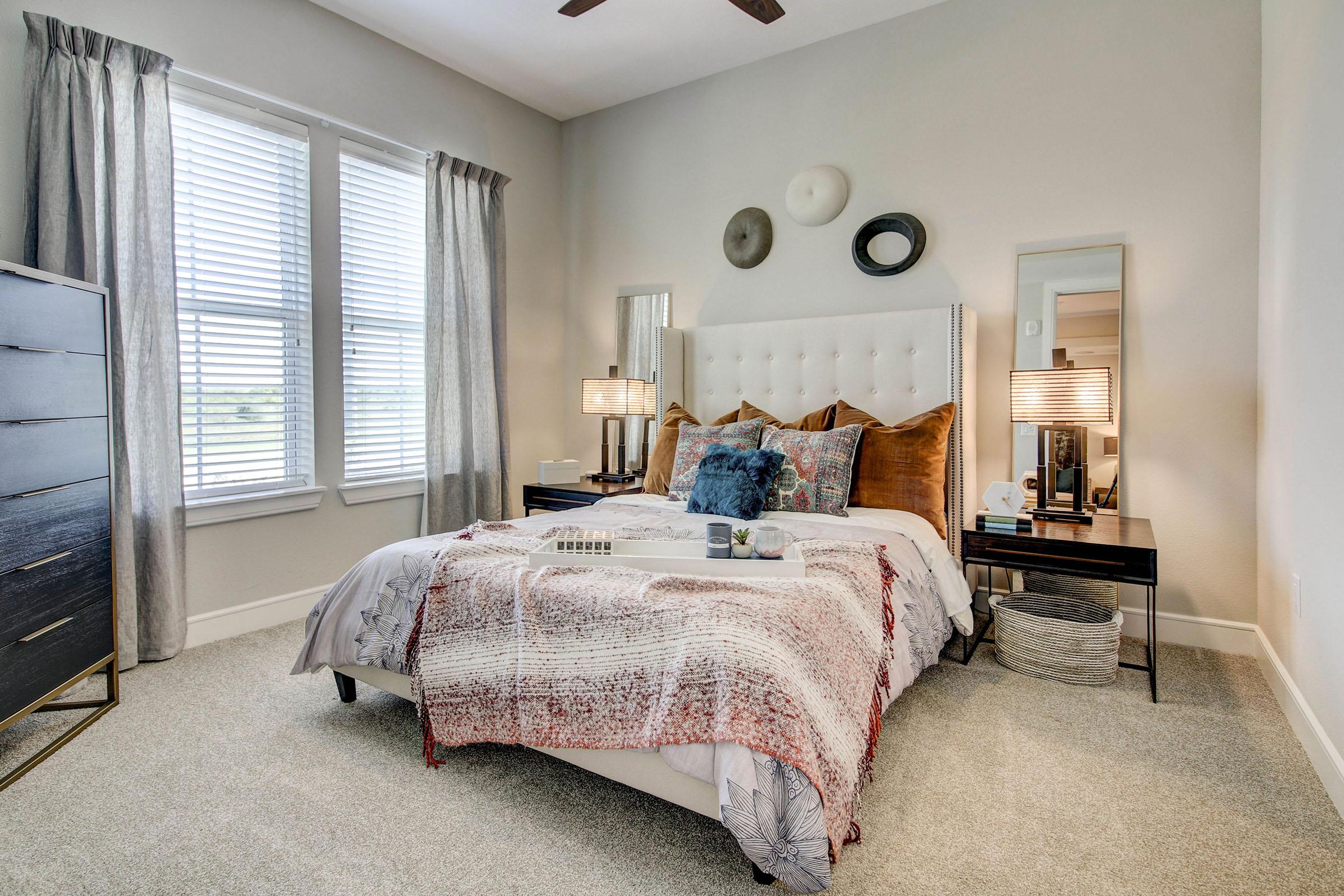 A cozy bedroom featuring a neatly made bed adorned with decorative pillows and a textured throw blanket. There are two bedside tables with stylish lamps, a tall dresser, and a wall with three decorative plates. Natural light filters in through two large window shades, enhancing the inviting atmosphere.
