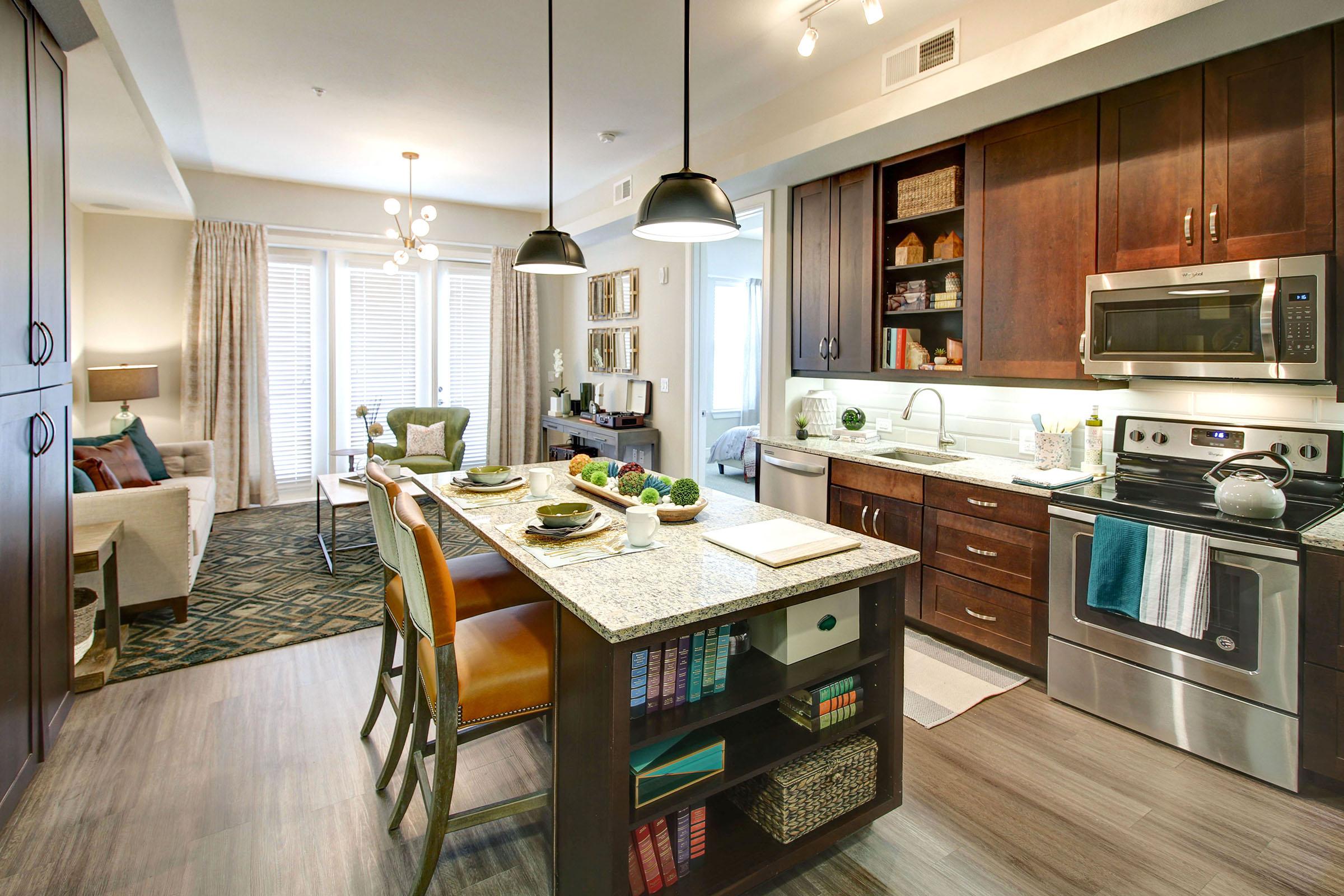 Modern kitchen and living area in an apartment. The kitchen features dark wood cabinets, stainless steel appliances, and a large island with seating. A cozy living space includes a sofa, decorative pillows, and large windows with natural light. The decor is stylish and inviting, with a blend of contemporary design elements.