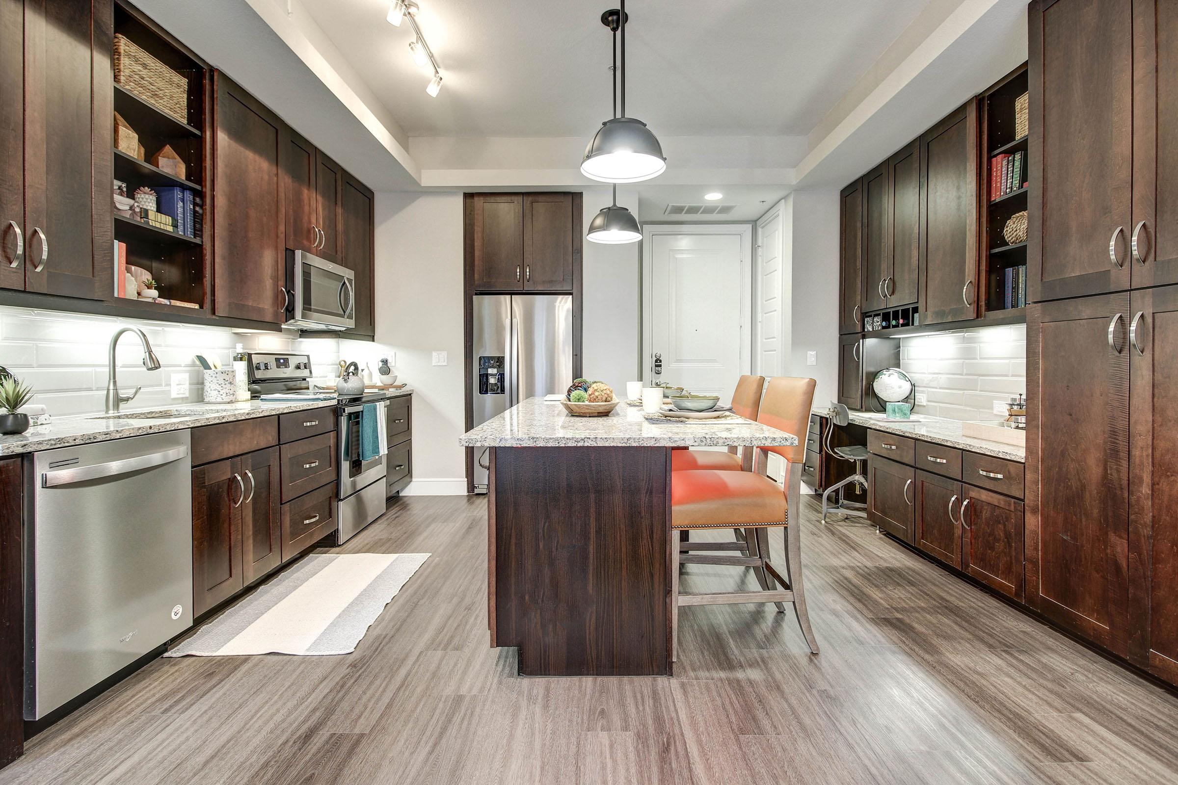 Modern kitchen featuring dark wood cabinetry, stainless steel appliances, and a spacious island with seating. The granite countertops complement the sleek design, while warm lighting creates an inviting atmosphere. A neatly arranged table with fruit adds a touch of decor.