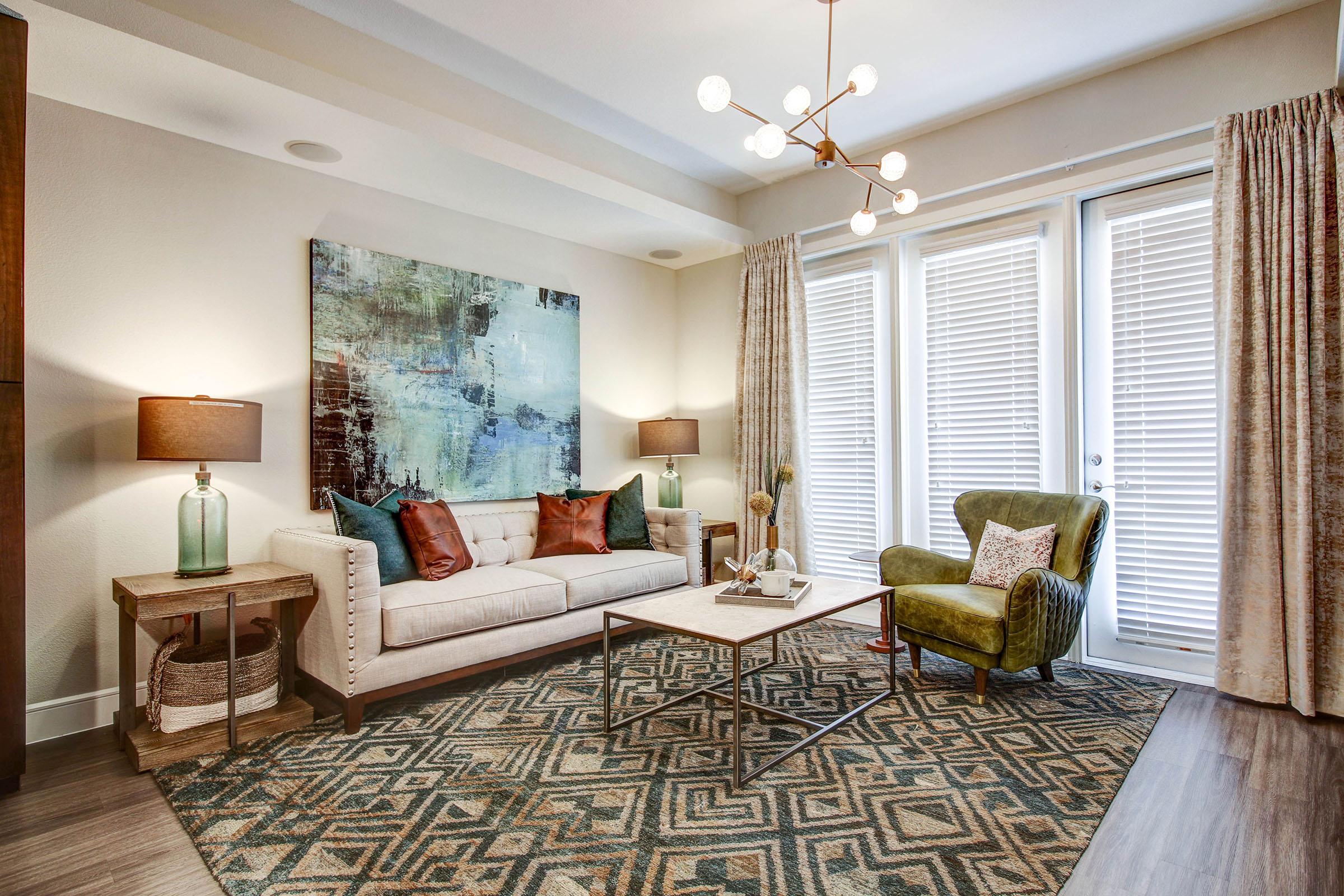 A modern living room featuring a light-colored sofa with patterned pillows, an abstract art piece on the wall, two stylish lamps on side tables, a glass coffee table, and a green armchair. Large windows with blinds let in natural light, complemented by curtains. A geometric rug adds texture to the space.