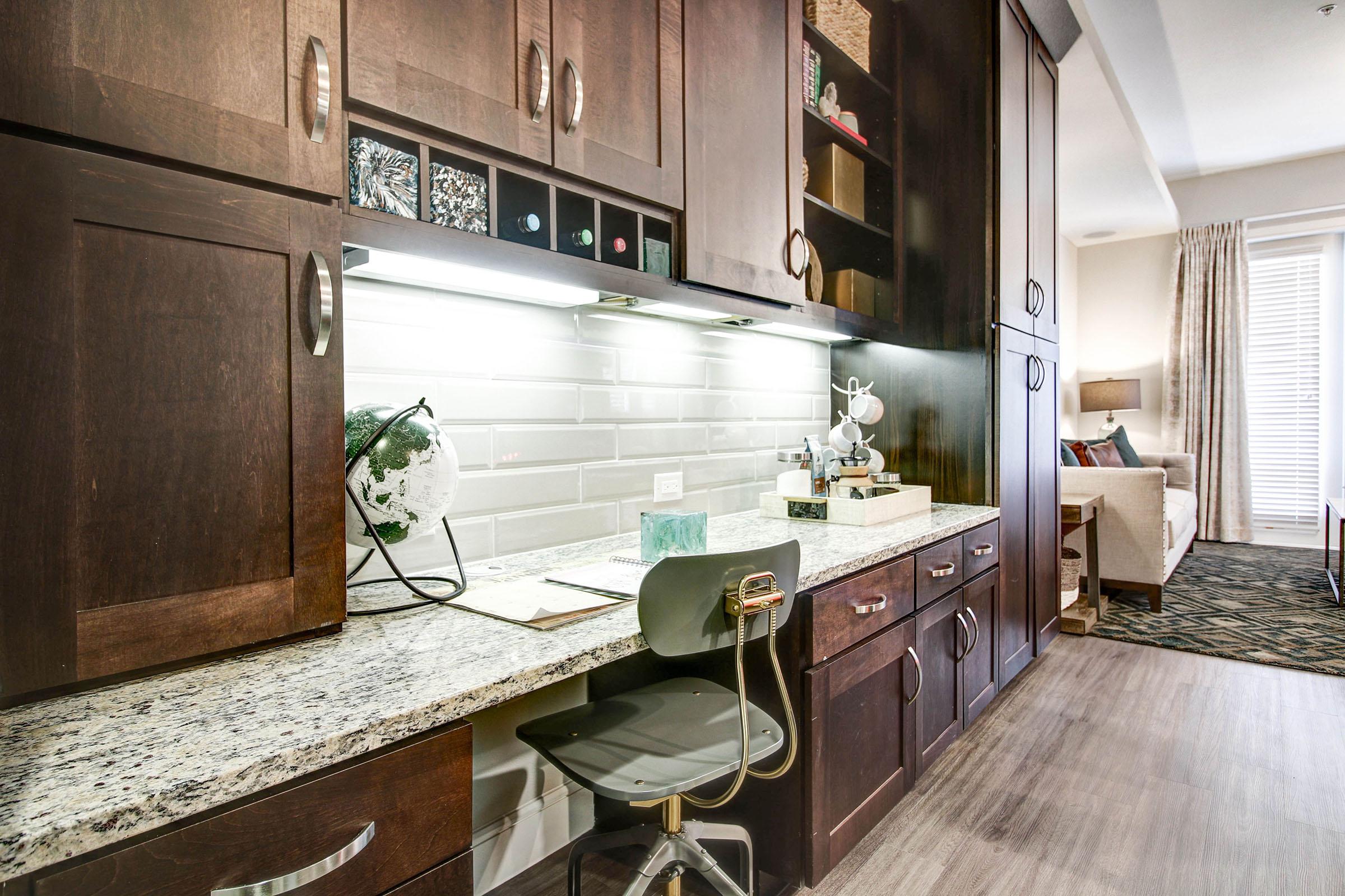 A stylish home office nook features dark wood cabinetry, a granite countertop, and modern decor. There’s a rolling chair, a globe, and neatly organized items on the shelves. Natural light floods the space from a nearby window, creating a cozy atmosphere.