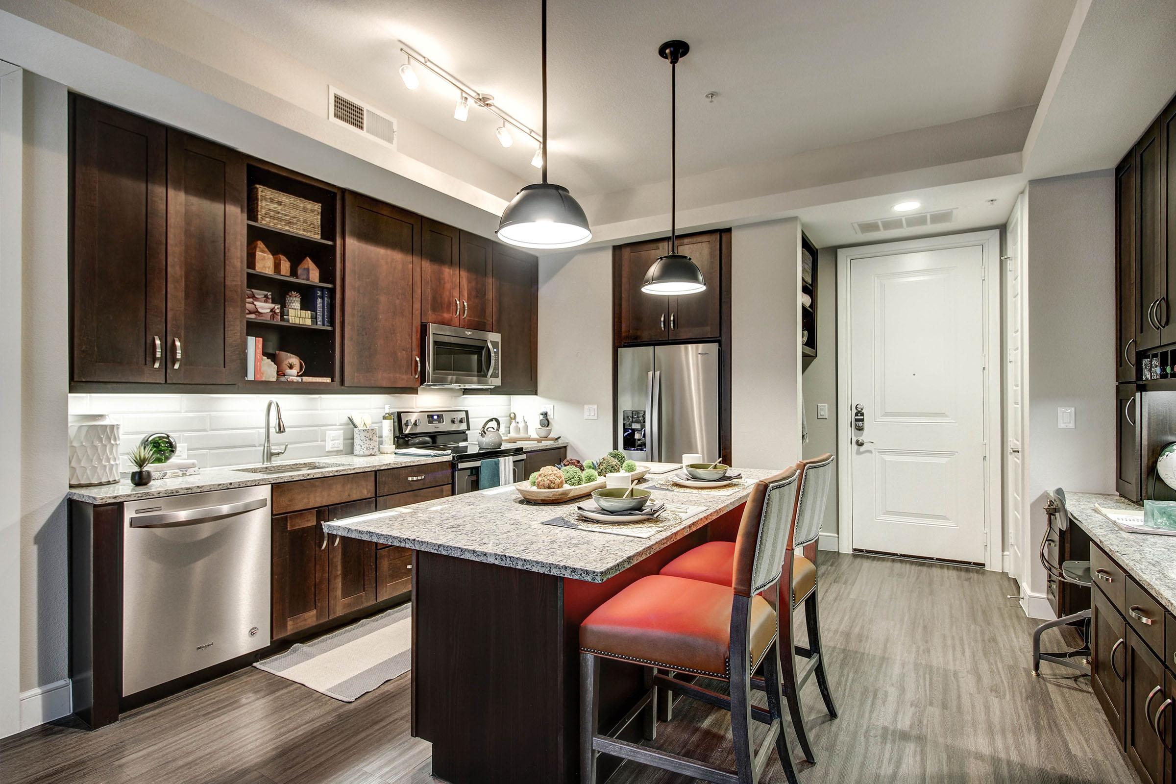 Modern kitchen featuring dark wood cabinetry, stainless steel appliances, and a large island with seating. Soft lighting accentuates the space, which includes dishes and decorative items on display. The entrance door is visible in the background, adding to the inviting atmosphere.