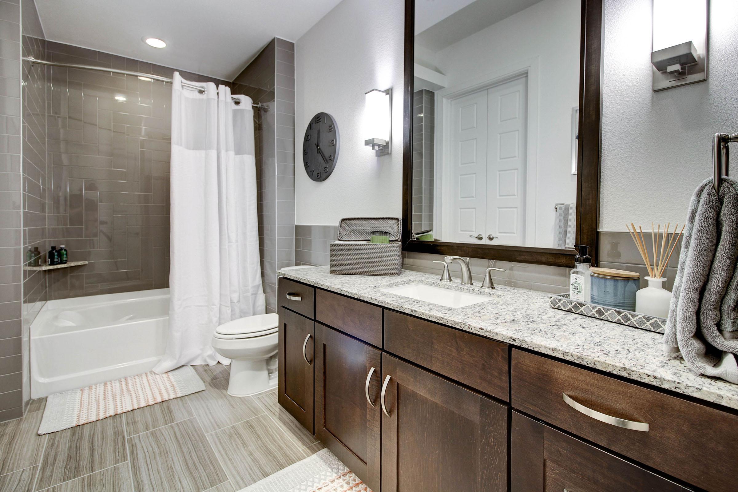 Modern bathroom featuring a bathtub with a shower curtain, a round wall clock, a double sink with a granite countertop, dark wooden cabinetry, and neatly arranged towels. The floor is tiled, and decorative items are visible on the countertop. Bright lighting enhances the overall ambiance.