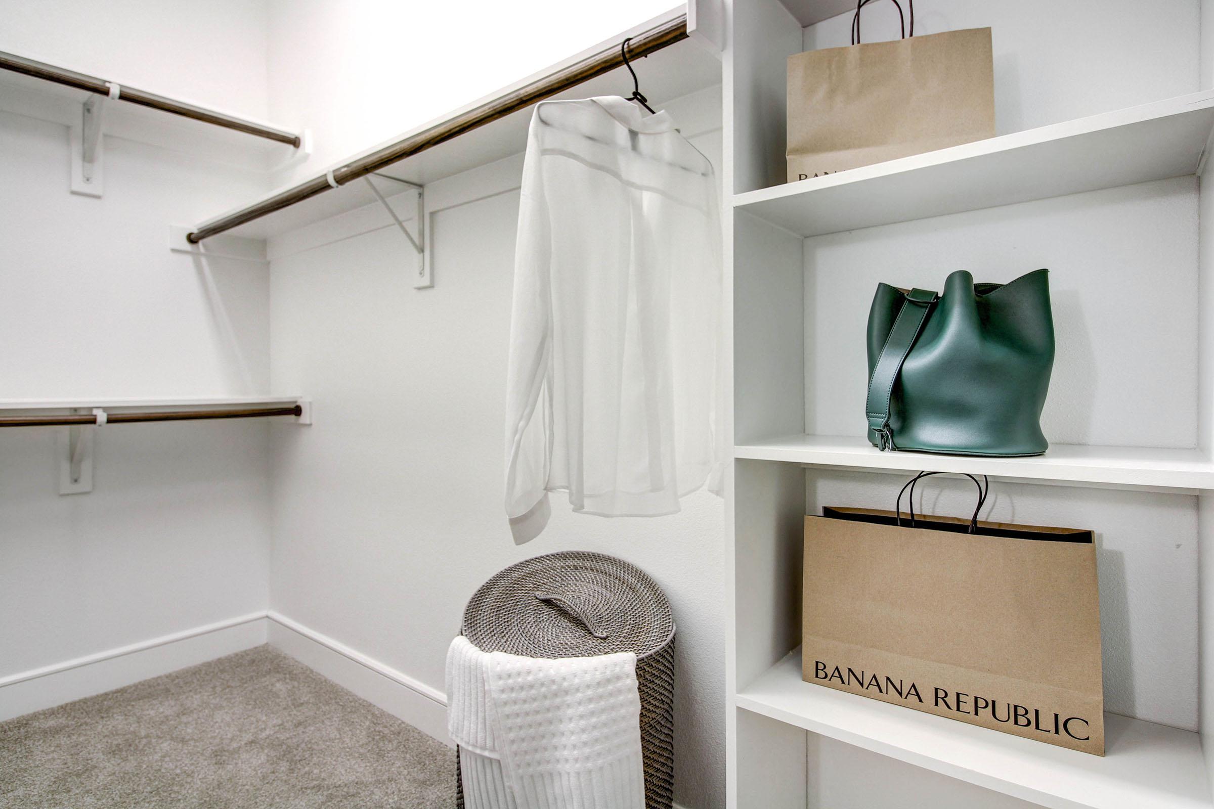 A spacious closet with a neatly hung white blouse, a woven basket, a green handbag on a shelf, and two shopping bags from Banana Republic. The walls are light-colored, and the floor has a plush, light gray carpet. The overall design is clean and organized, showcasing a modern aesthetic.