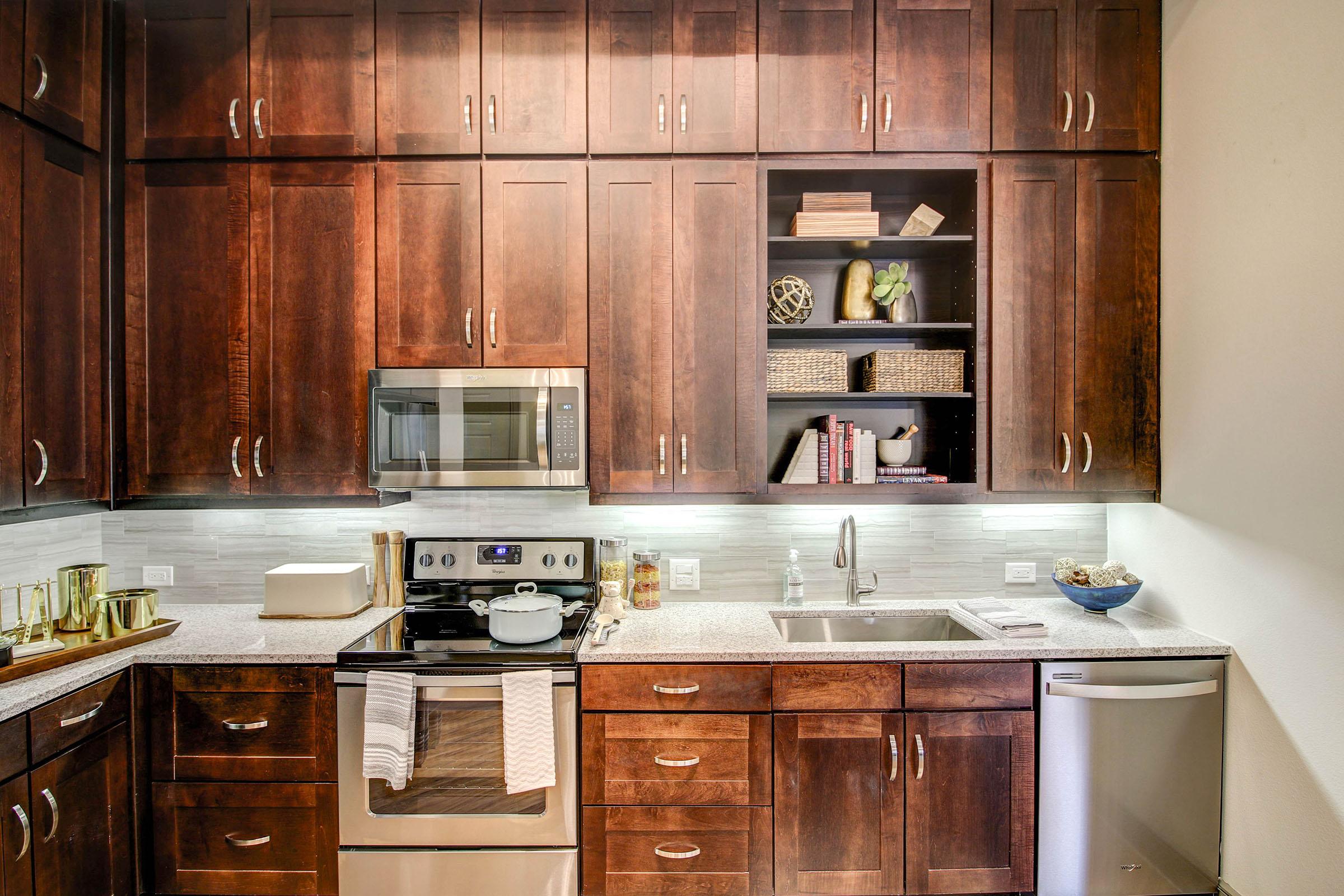 A modern kitchen featuring dark wood cabinetry, a stainless steel stove and microwave, a white countertop, and a stylish backsplash. Shelves display decorative items and storage baskets, while a small fridge is visible in the corner. The overall design is sleek and contemporary.