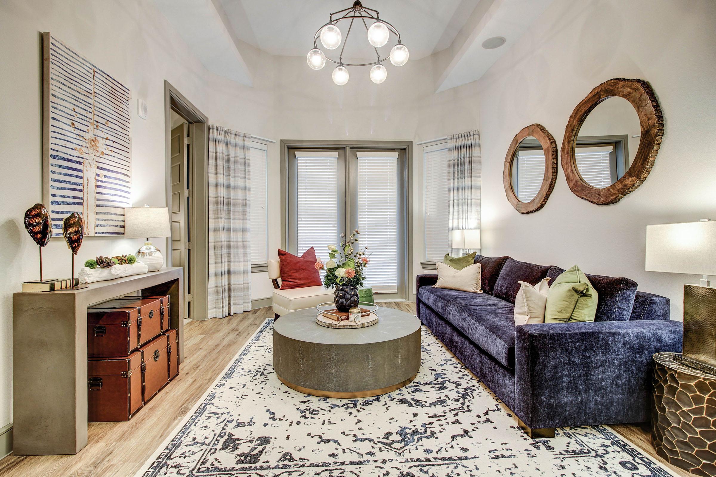 A stylish living room featuring a dark blue velvet sofa, a round coffee table, and a light-colored accent chair. Decorative items include a floral arrangement on the table, two mirrors on the walls, and lamps on side tables. The floor is covered with a patterned area rug, and natural light filters through window treatments.