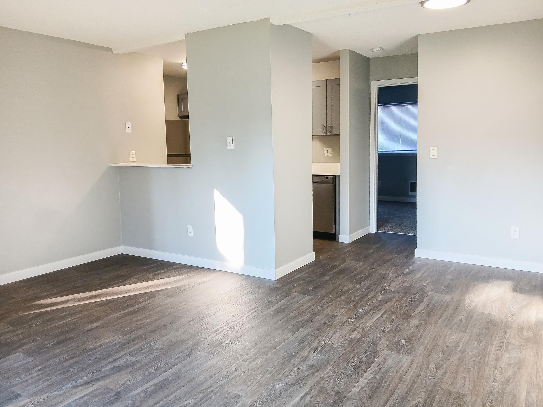 An interior view of a modern, empty living space featuring light gray walls and wood-like flooring. Natural light streams in from a window, illuminating the room. A kitchenette can be seen in the background, and an open doorway leads to another room. The atmosphere is bright and inviting.