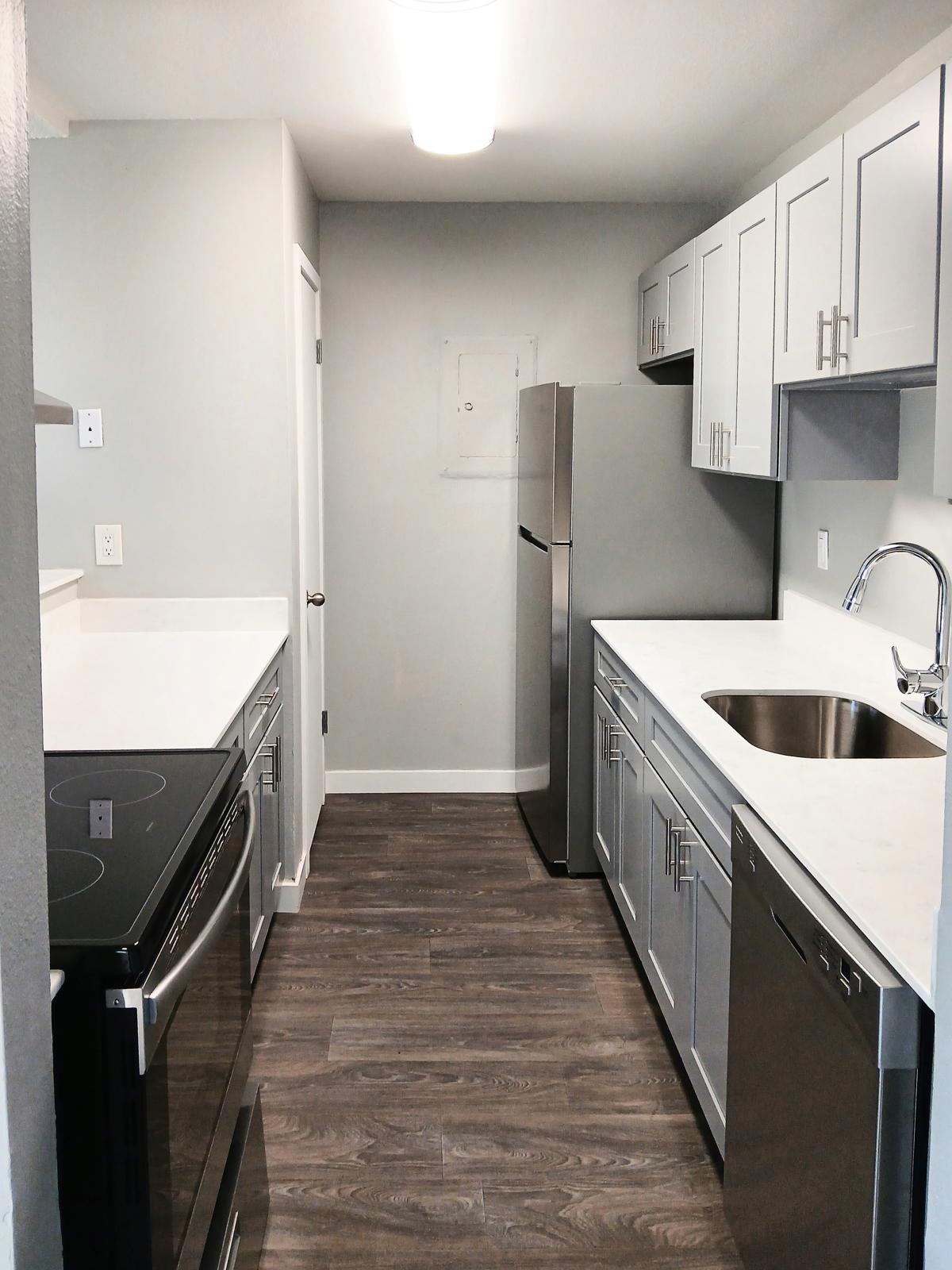 A modern kitchen featuring sleek, gray cabinetry, a stainless steel refrigerator, a built-in oven, and a dishwasher. The countertops are light-colored, and the space has wood-like flooring. A single basin sink is installed next to the dishwasher, with bright overhead lighting illuminating the area.