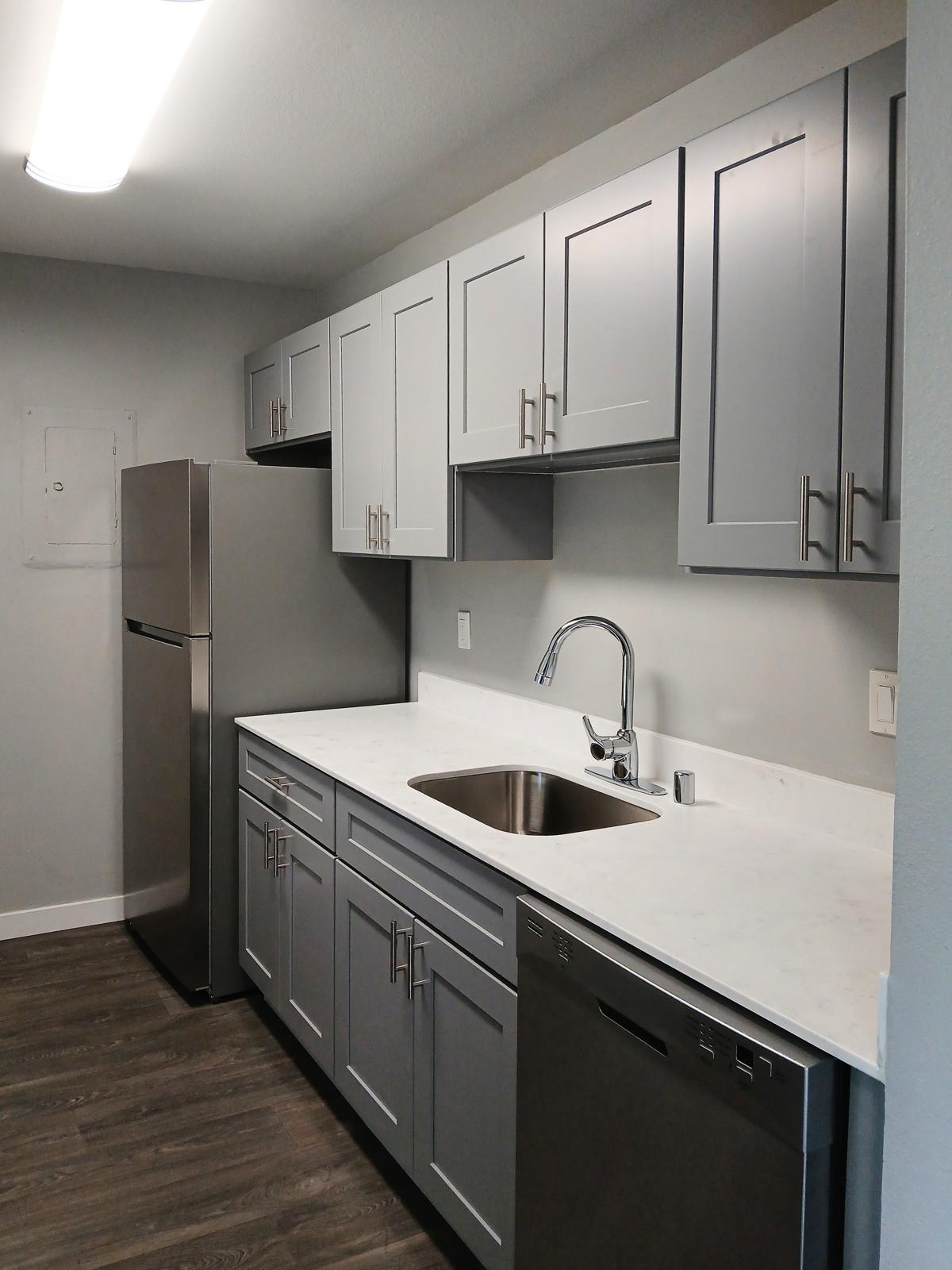 Modern kitchen featuring gray cabinets, a stainless steel refrigerator, a double sink with a sleek faucet, and a dishwasher. The counter is made of white marble, and the flooring is dark wood, creating a stylish and functional space with plenty of storage.