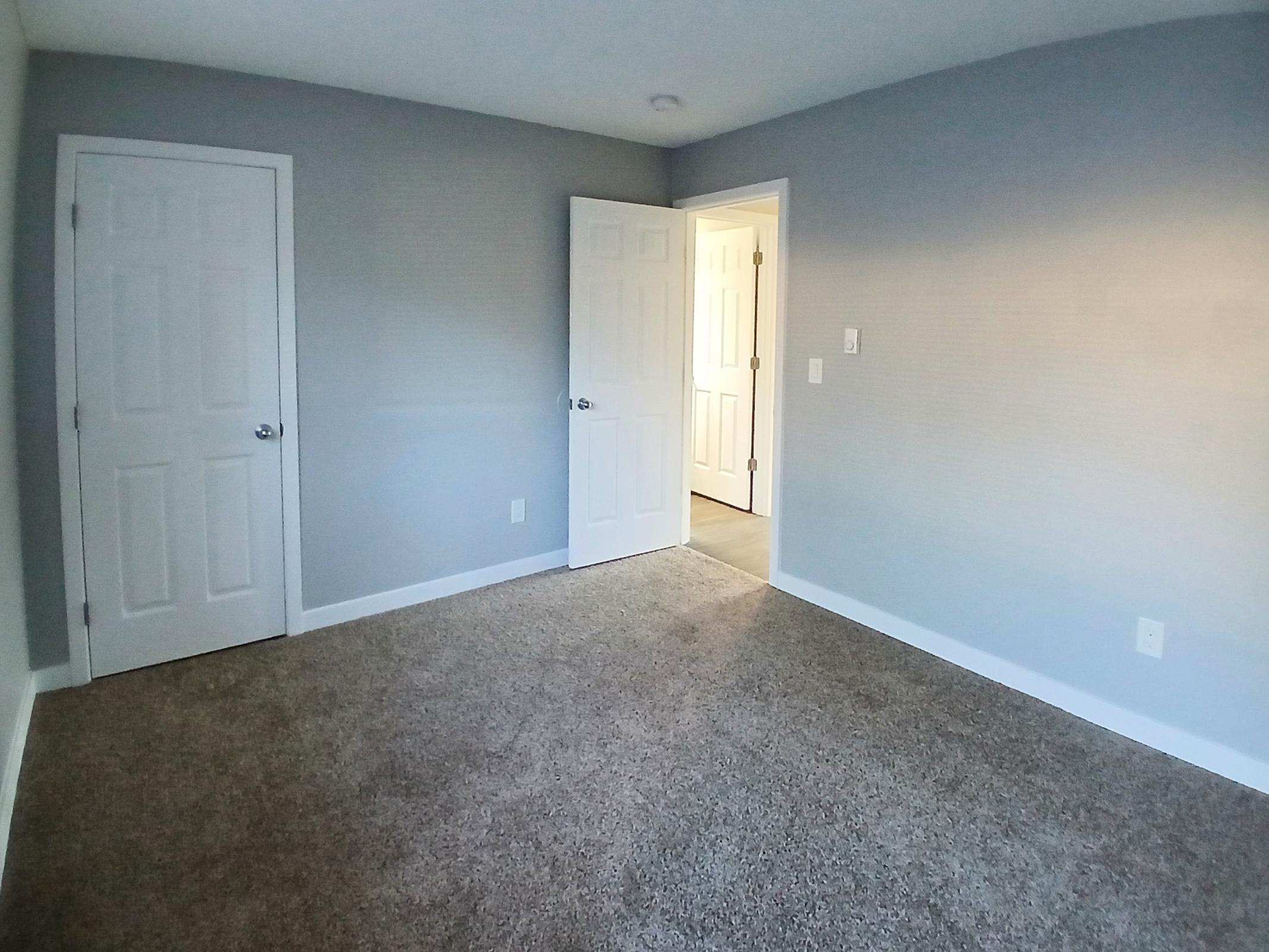 A spacious, empty room with light gray walls and carpeted flooring. Two white doors are visible, one slightly ajar, leading to another room. Natural light filters in from the open doorway, illuminating the subtle texture of the walls. The space appears clean and inviting.