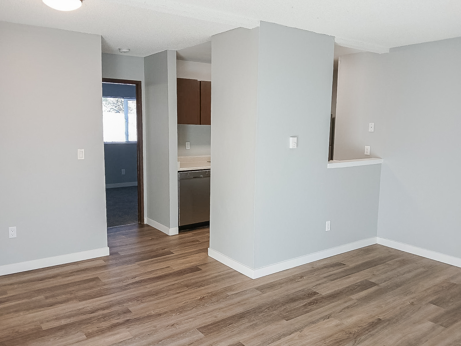 A spacious, well-lit room featuring light gray walls and laminate flooring. On the left, there's a doorway leading to another room, while the kitchen area is partially visible with wooden cabinets and a stainless steel dishwasher. The overall design is modern and inviting.