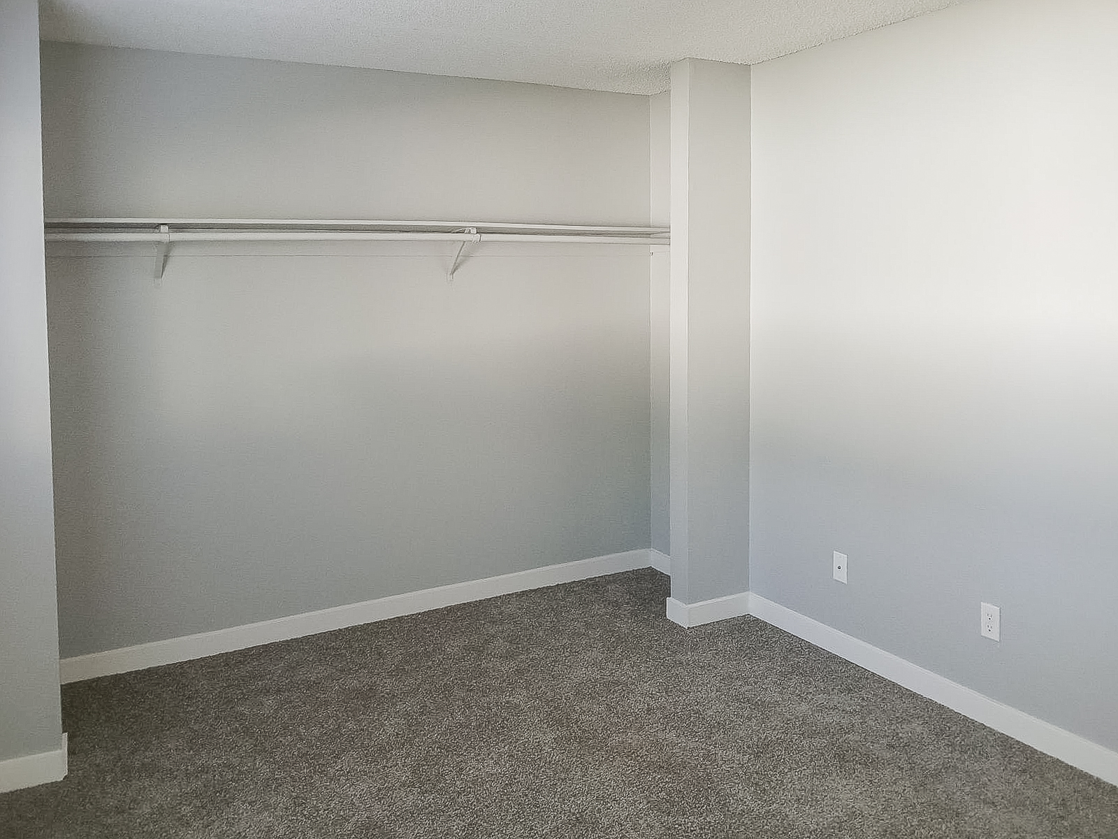 Empty room with light gray walls and carpeted floor. There's a built-in shelf along one wall, and the space is well-lit, suggesting it could serve as a closet or small storage area. No furniture or decorations are present, giving it a minimalist appearance.