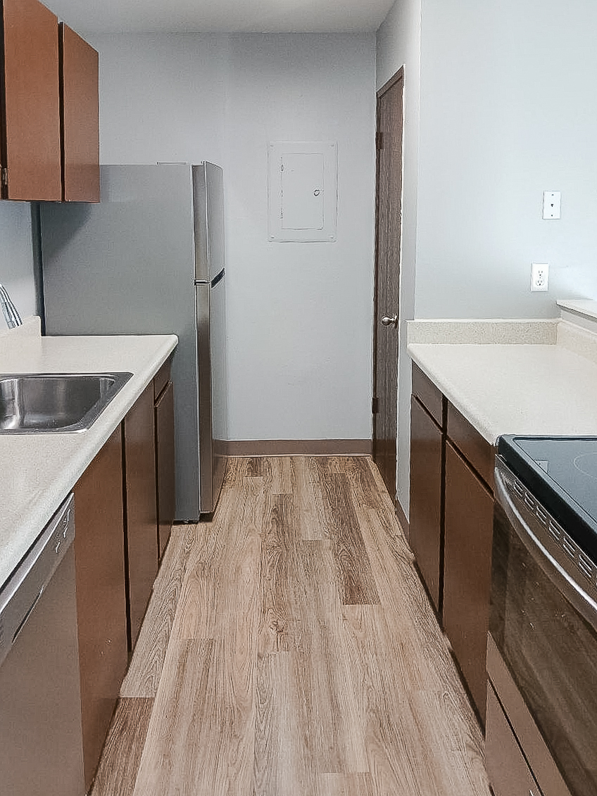 A modern kitchen featuring wood cabinets, stainless steel appliances including a refrigerator and stove, a sink with a countertop, and light-colored walls. The floor is made of wood laminate, and there is a door leading to another room visible at the end of the kitchen.