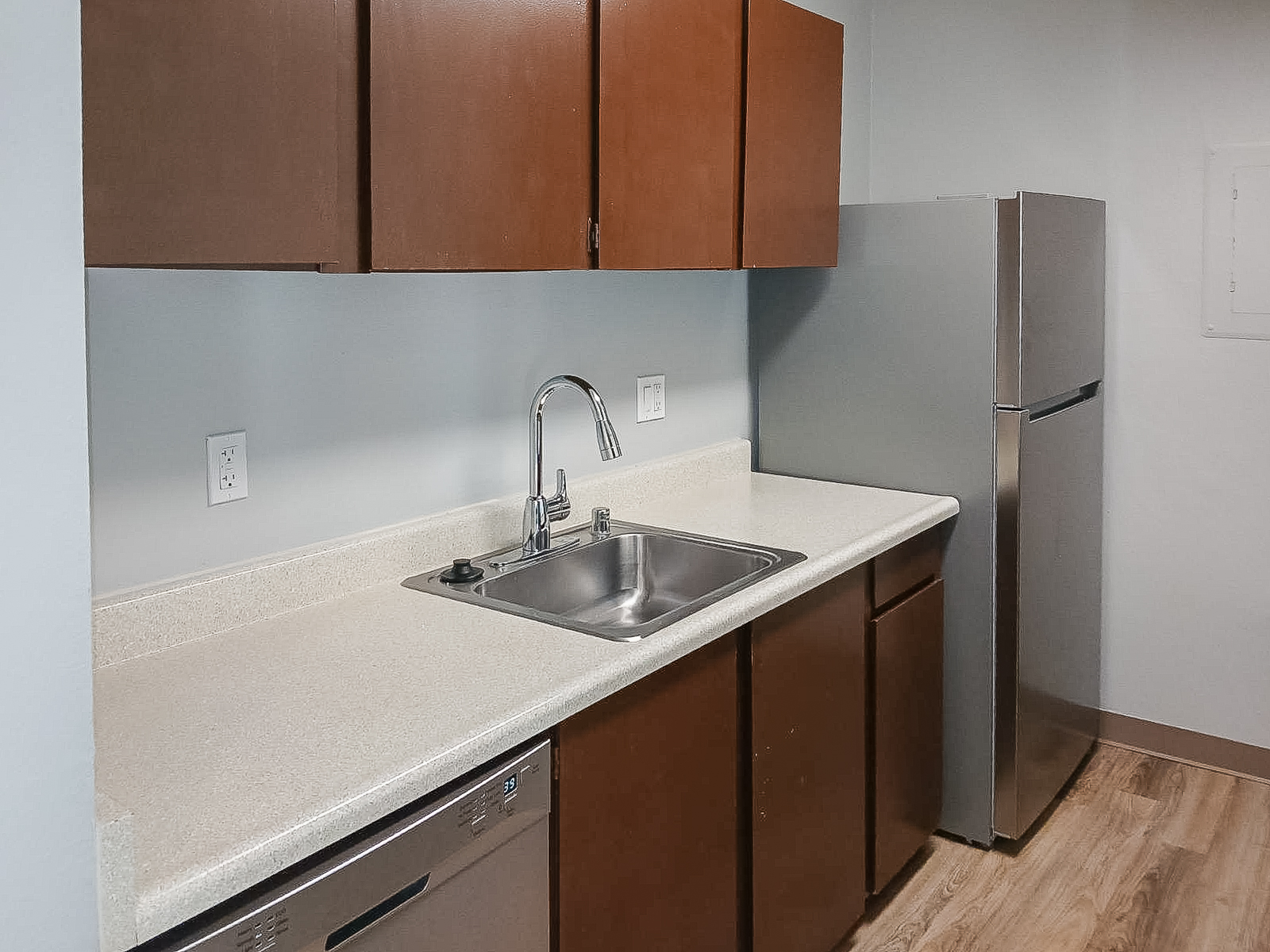 A modern kitchen featuring a stainless steel refrigerator, a stainless steel sink with a single faucet, and dark wooden cabinetry. The countertop is light-colored, and the walls are painted in a soft blue. The flooring is a light wood finish, creating a clean and contemporary look.