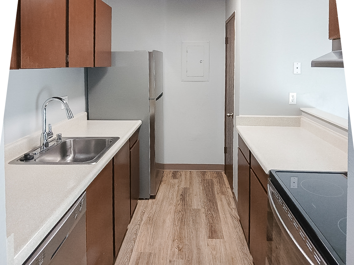 A modern kitchen featuring light-colored cabinets, stainless steel appliances, and a double sink. The flooring is a light wood laminate, and the countertops are a neutral color. There is a refrigerator, a dishwasher, and an electric stove, with ample workspace and wall-mounted electrical outlets.