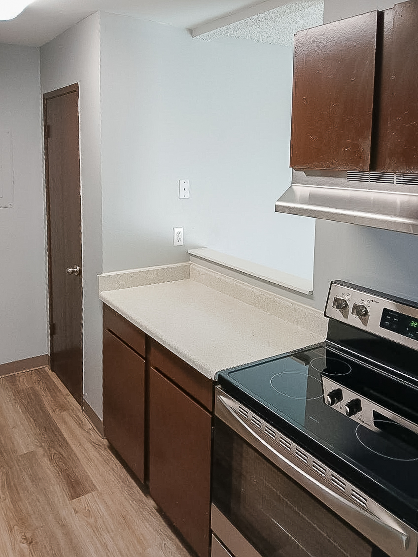 A modern kitchen featuring a countertop made of light-colored material, wooden cabinets, and a stainless steel oven and stove. The space has neutral-colored walls and a door leading to another room. The kitchen has a clean and minimalistic design, with a simple layout.