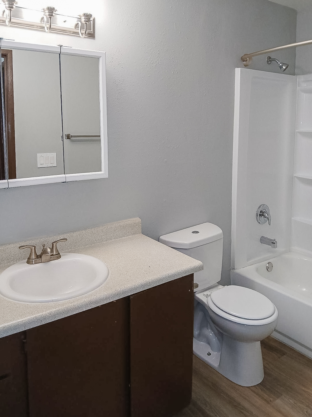 A clean bathroom featuring a countertop with a sink, a wall-mounted mirror, and a toilet. There is a bathtub with a shower curtain and a showerhead. The walls are painted in a light gray color, and the flooring is a wood-like laminate. Soft lighting illuminates the space from a fixture above the mirror.