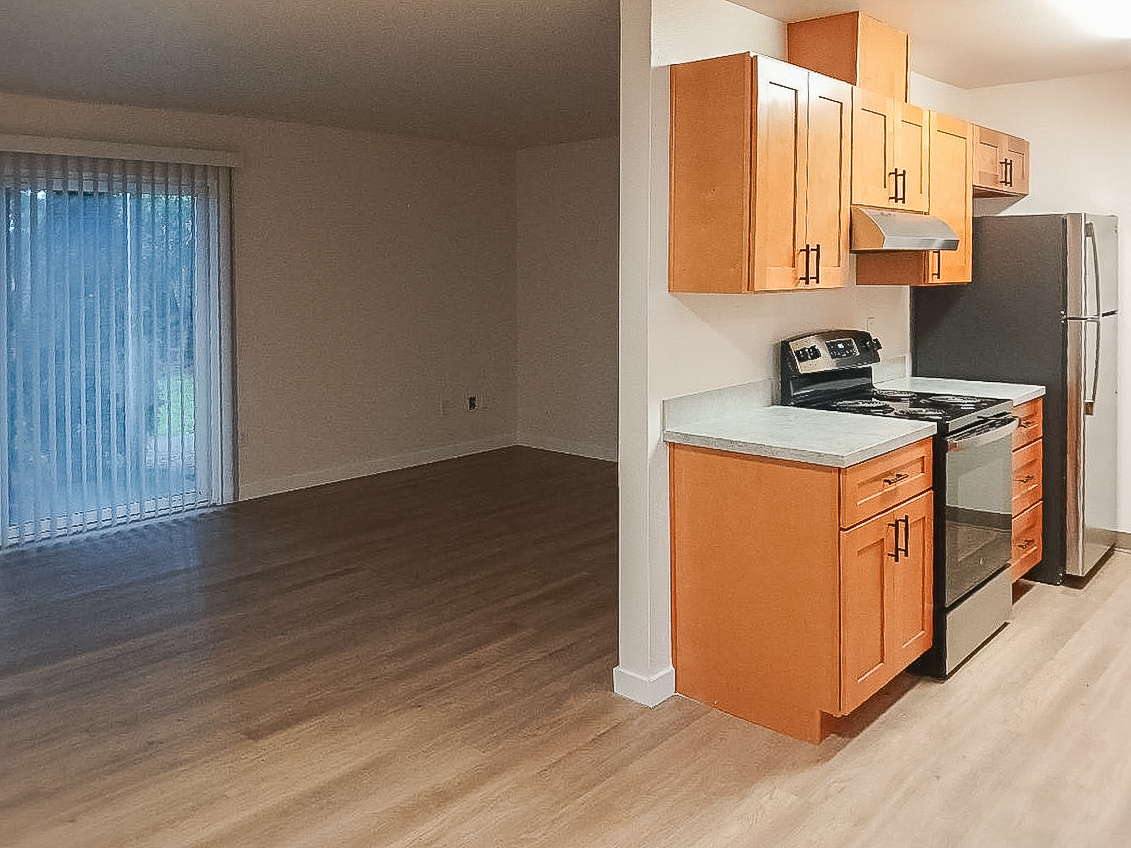 A modern kitchen featuring wooden cabinets, a stainless steel stove, and a refrigerator, with a gray countertop. Adjacent to the kitchen is a spacious living area with light-colored wooden flooring and a sliding glass door leading to an exterior space. The overall atmosphere is bright and inviting.