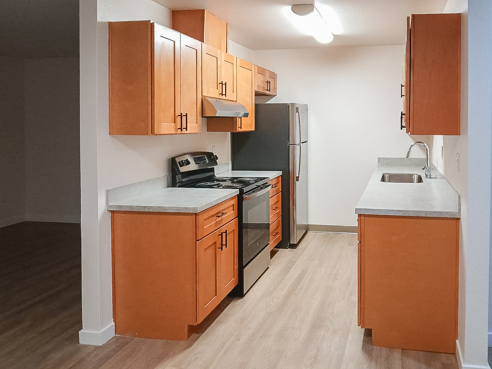 A modern kitchen featuring wooden cabinets with a light orange finish, stainless steel appliances including a stove and refrigerator, gray countertops, and a sink. The space has a neutral-colored wall and a wood-look floor, creating a warm and inviting atmosphere.