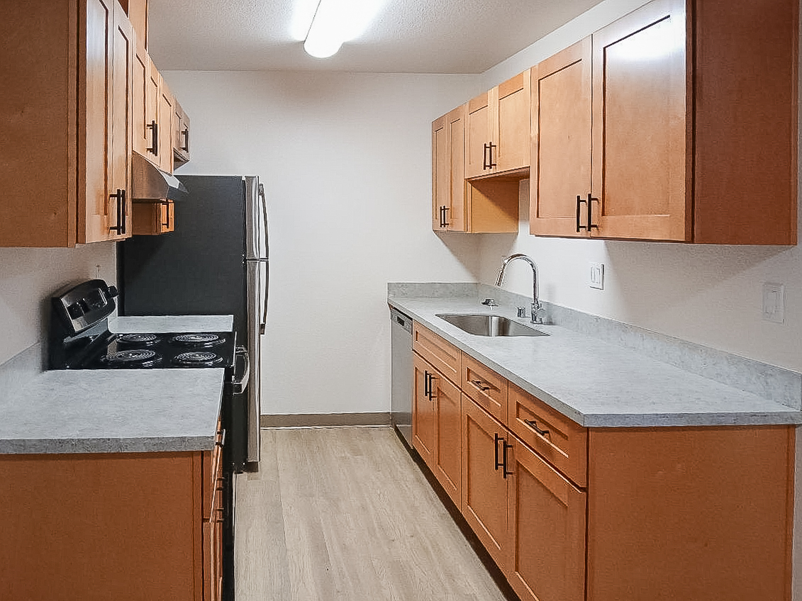 Modern kitchen featuring wooden cabinets with a light finish, gray countertops, a stainless steel refrigerator, an electric stove with an oven, and a sink. The space is well-lit with overhead fluorescent lighting and has a simple, clean layout.