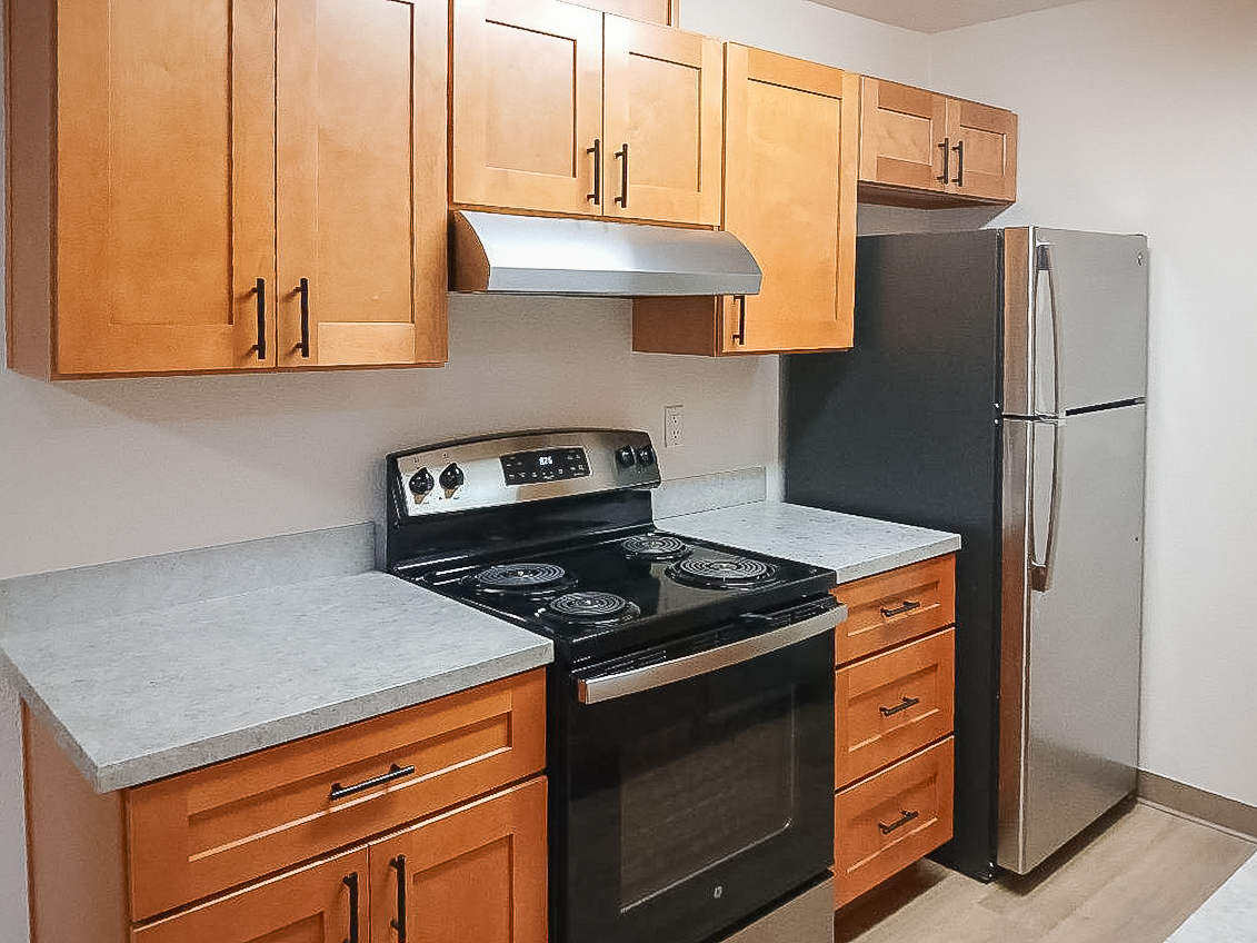 A modern kitchen featuring wooden cabinetry, a silver refrigerator, and a black stove with an oven. The countertops are light gray, and there is a stainless steel range hood above the stove. The kitchen has a clean, contemporary look with ample storage space and appliances.