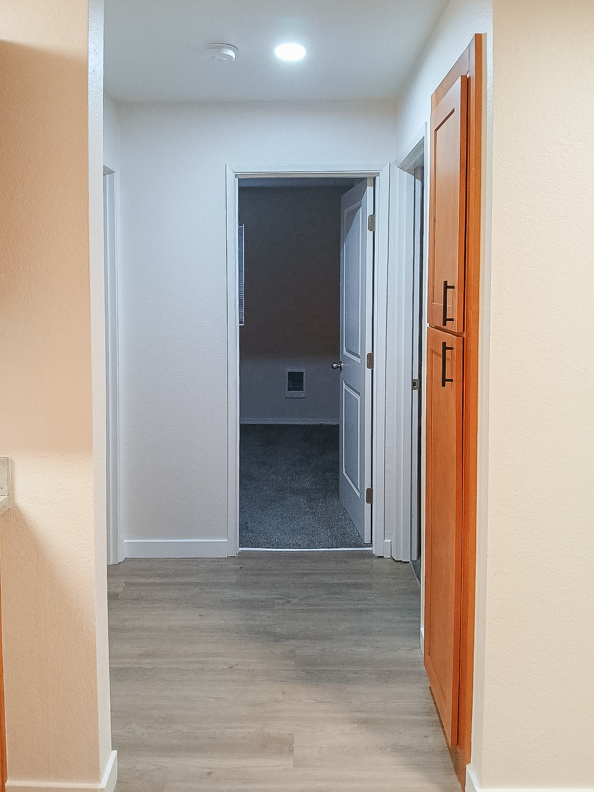 A narrow hallway with light beige walls and wooden trim. On the left, there are closed doors and a tall cabinet. At the end of the hallway, an open doorway leads to a room with dark carpeting. The ceiling is bright with recessed lighting.