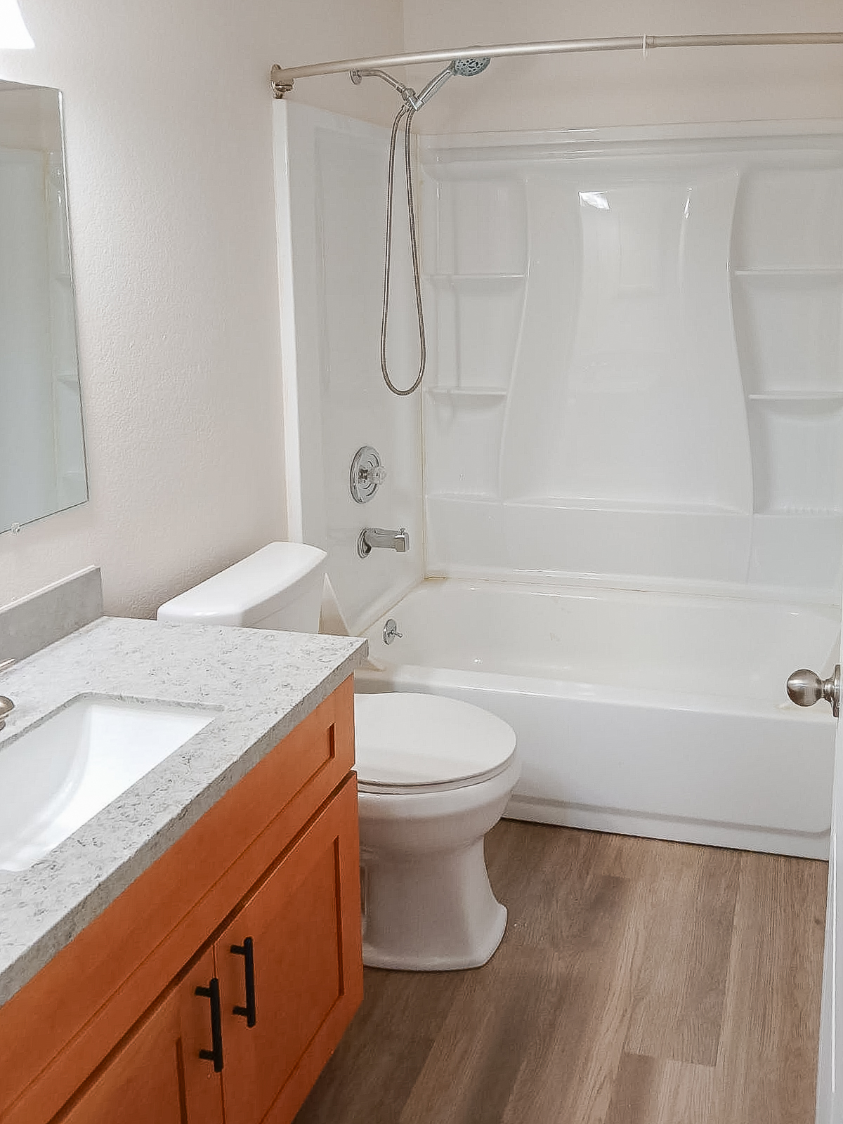 A clean and modern bathroom featuring a white bathtub and shower, a mirror above a sink with a granite countertop, and light wooden cabinetry. The floor is made of light-colored wood-like tiles, creating a welcoming and spacious atmosphere.