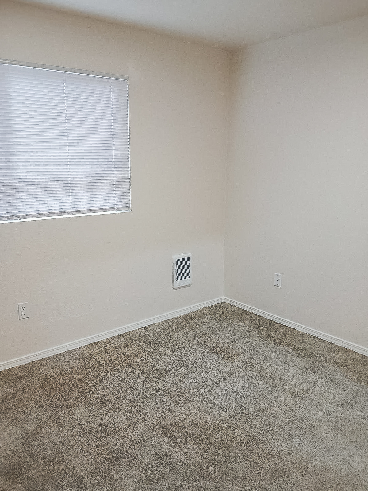 Empty room with light beige walls and a window covered by white blinds. The floor is covered in light brown carpet, and there is a small vent on one wall. Minimal furnishings create an open and spacious feel.