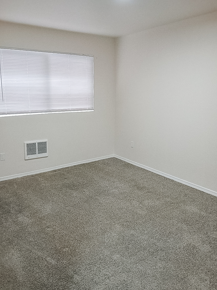 A vacant, unfurnished room with light-colored walls and a window featuring blinds. The floor is covered in light gray carpet, and there's a small vent on the wall near the floor. The room appears bright and spacious, with a neutral color scheme.