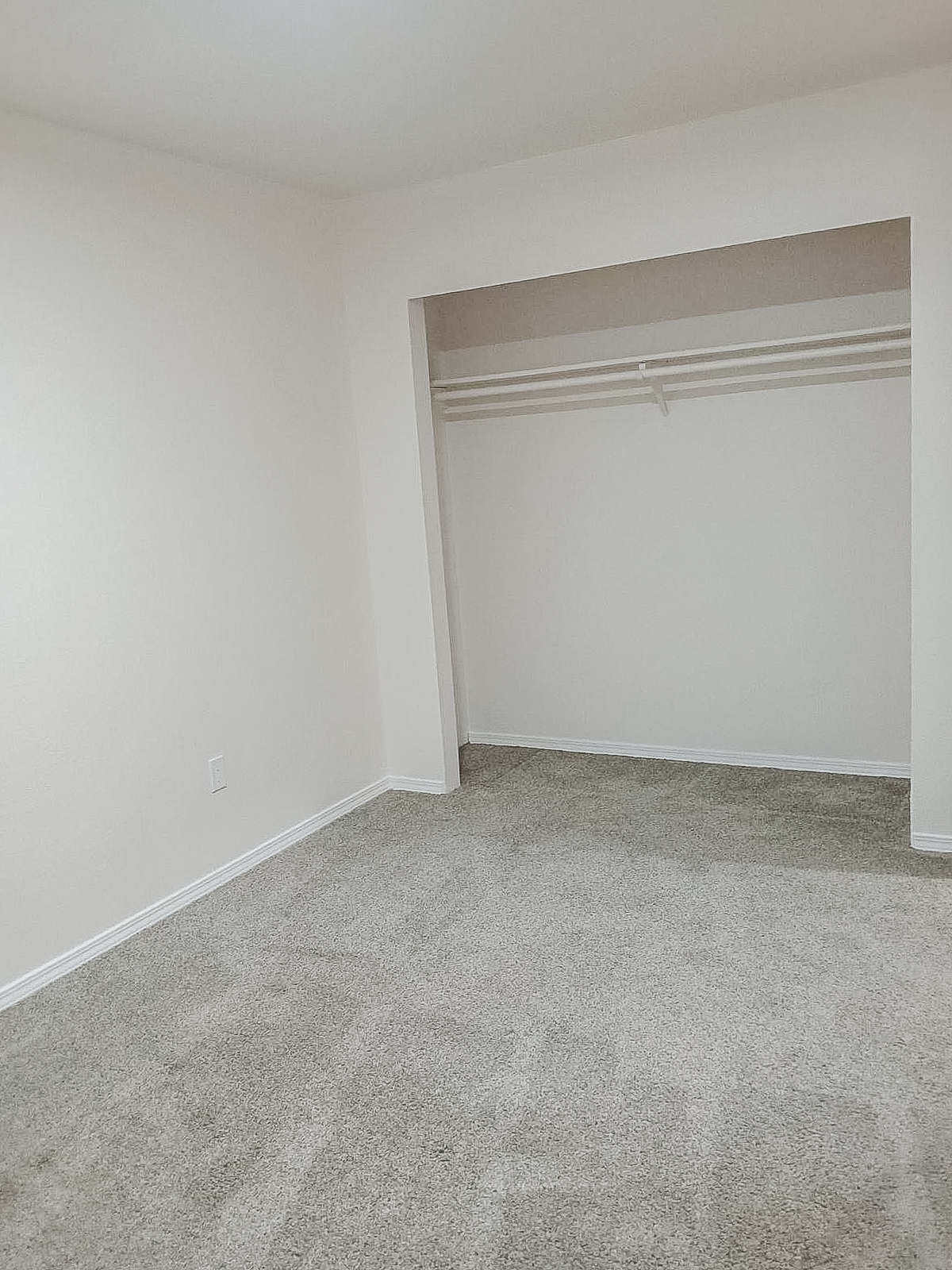 A spacious, empty room with light-colored walls and beige carpet. There is a small alcove with a closet featuring a single hanging rod, and the overall look is minimalist and clean, suitable for various uses.