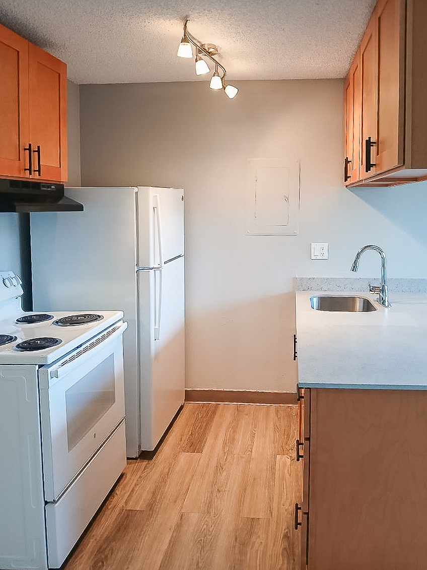 A kitchen featuring wooden cabinets, a white refrigerator, a white stove with a glass top, and a stainless steel sink. The backsplash is simple, and the floor has a light wood finish. The room is well-lit with overhead lighting, creating a warm and inviting atmosphere.