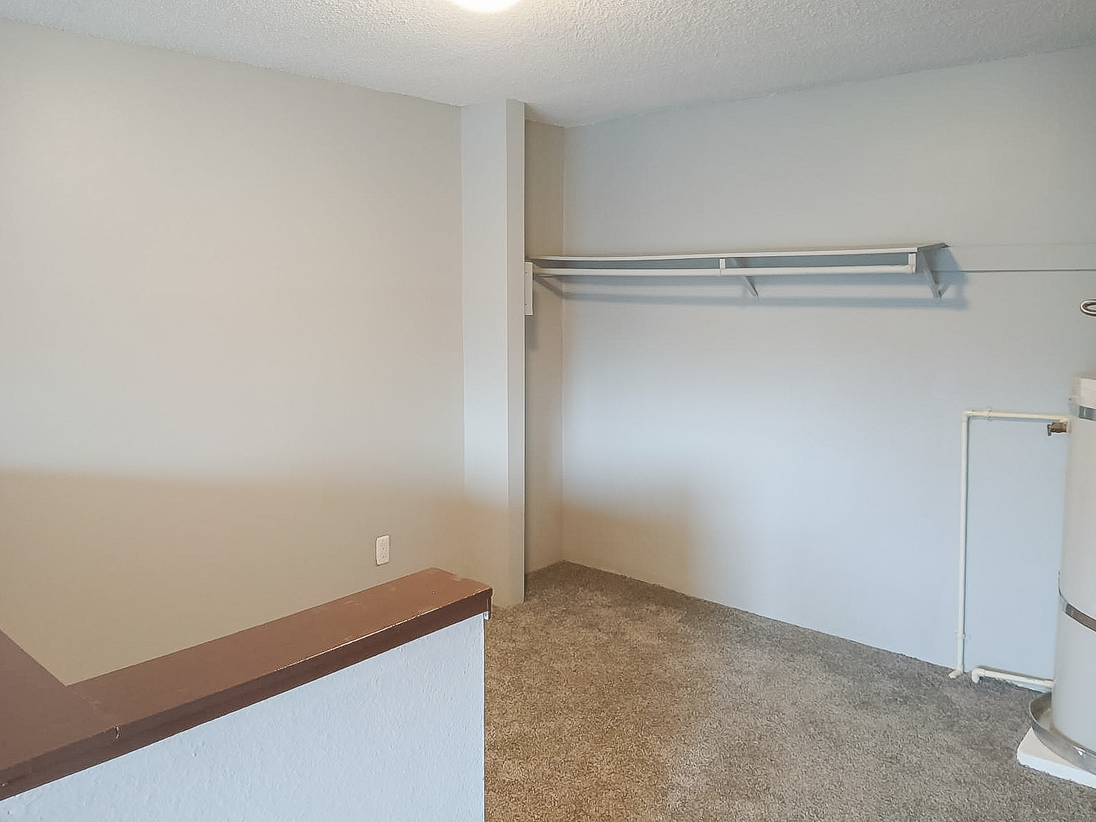 A small, empty room with light gray walls and a carpeted floor. There is a wall shelf on the left side, and a water heater is partially visible on the right. Natural light enters from an unseen source, creating a bright, airy atmosphere. The space appears suitable for storage or as a small utility area.