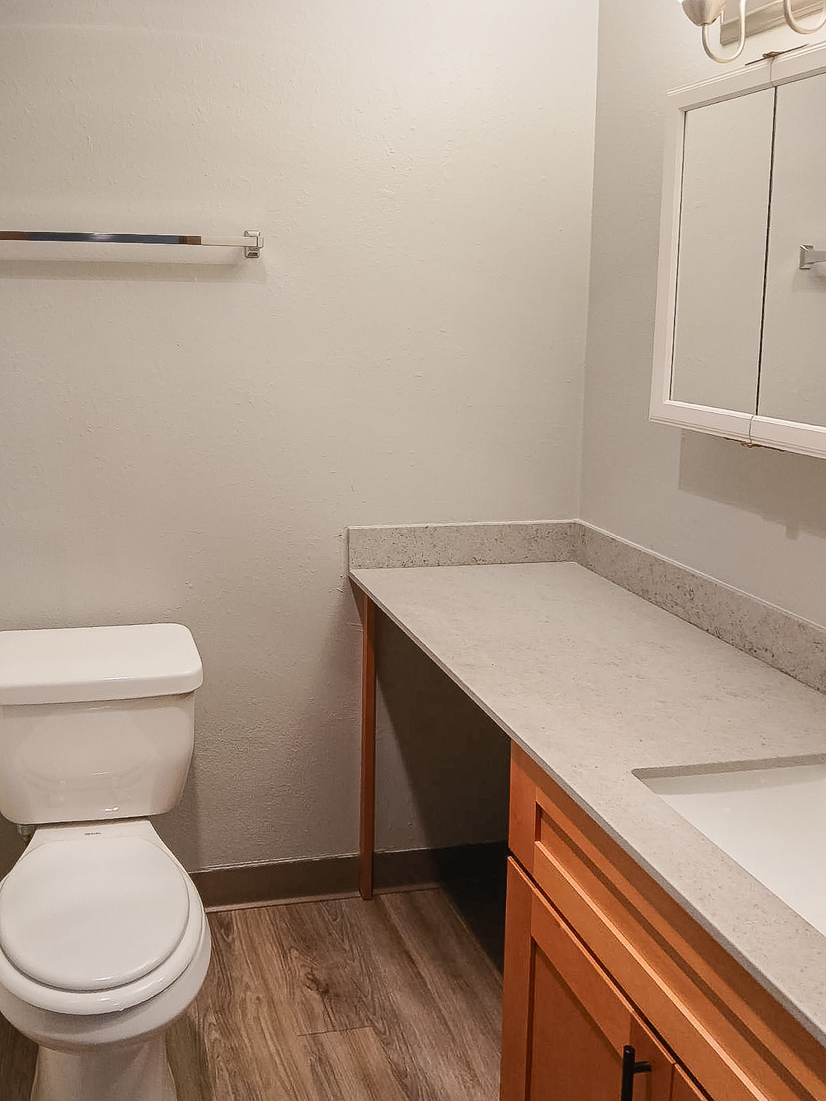 A clean bathroom interior featuring a modern toilet, a countertop with a sink, and a large mirror above the sink. The walls are painted light gray, and there is a small shelf mounted on the wall. The flooring is made of light wood or laminate.